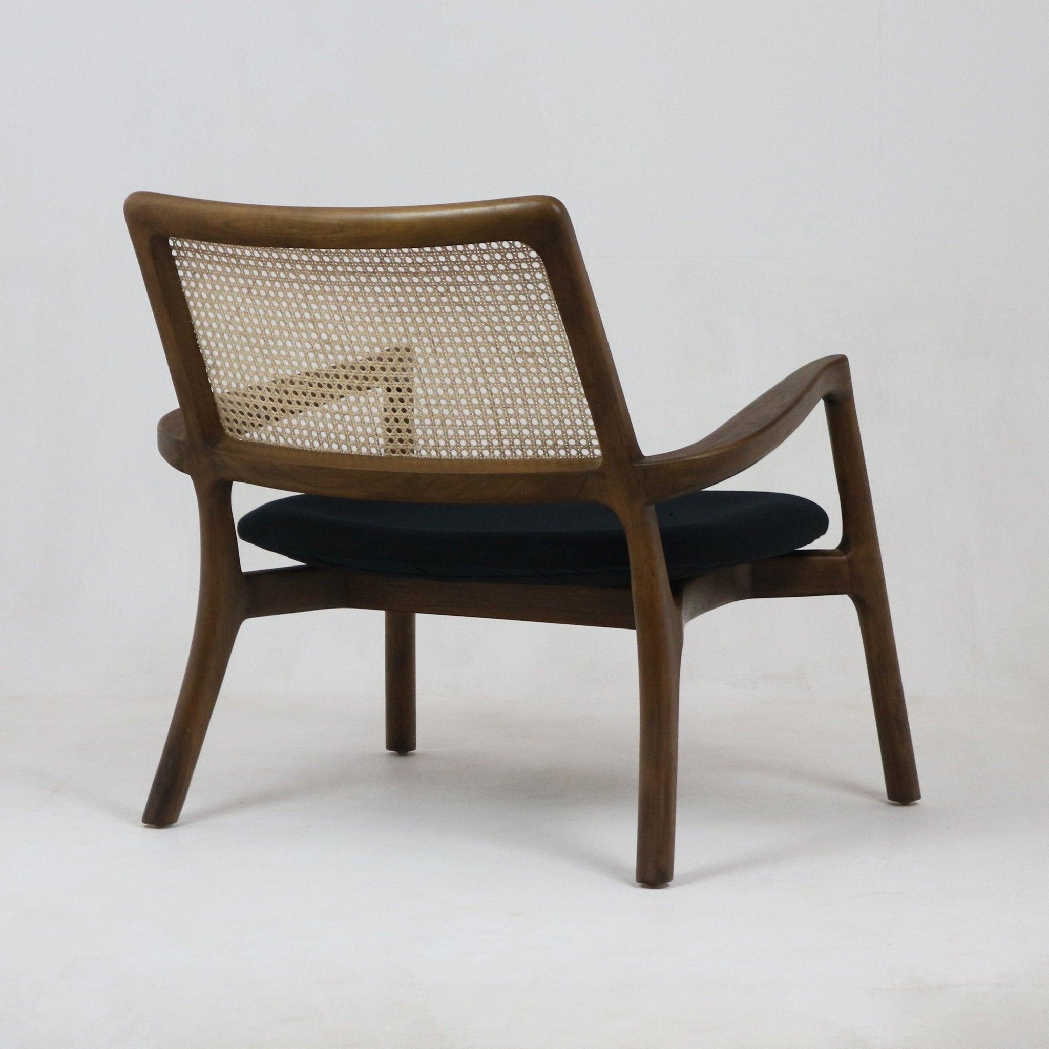Rear three-quarter view of a mid-century modern teak lounge chair featuring a woven cane back, sculpted teak frame with curved armrests and a dark fabric seat cushion.
