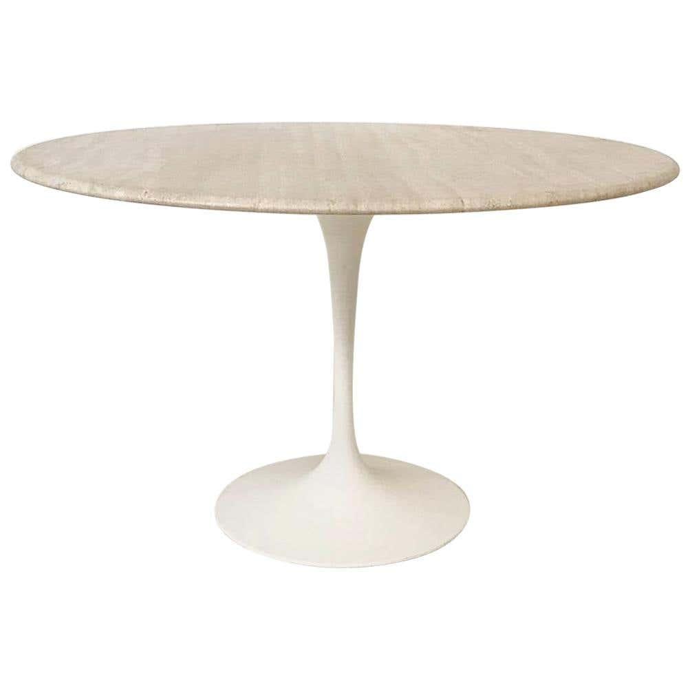 Round outdoor dining table with a beige travertine stone top and white metal tulip pedestal base in a modern minimalist style.