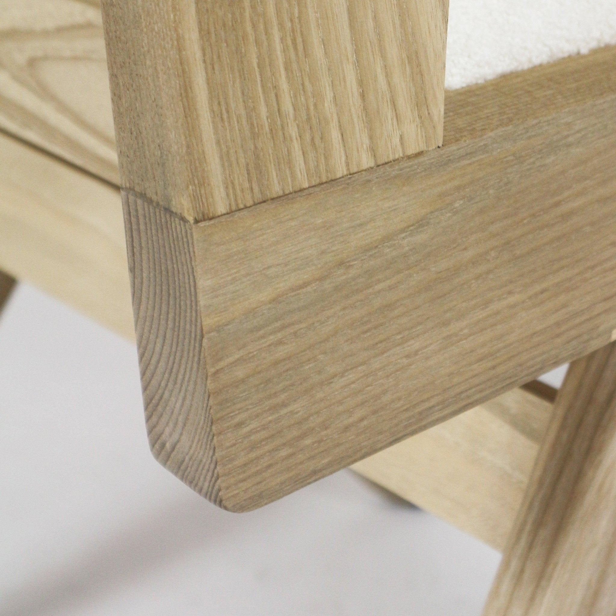 Close-up of a natural blonde solid ash chair frame corner and rounded end showing oak-like wood grain and a peek of cream boucle upholstery on the seat back.
