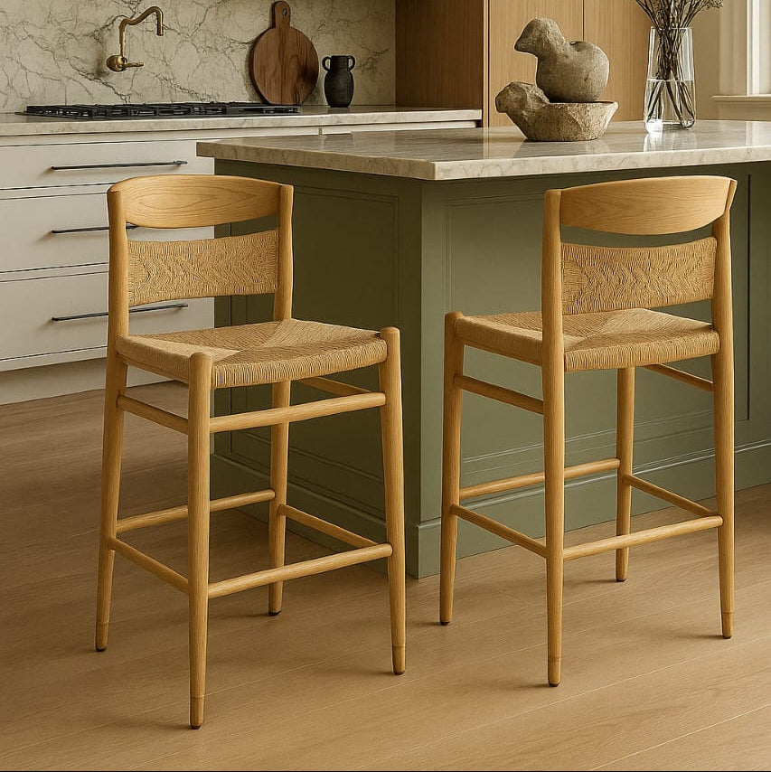 Two natural solid wood mid-century modern bar stools with woven papercord seats and curved wooden backrests placed at a green kitchen island topped with a marble countertop.