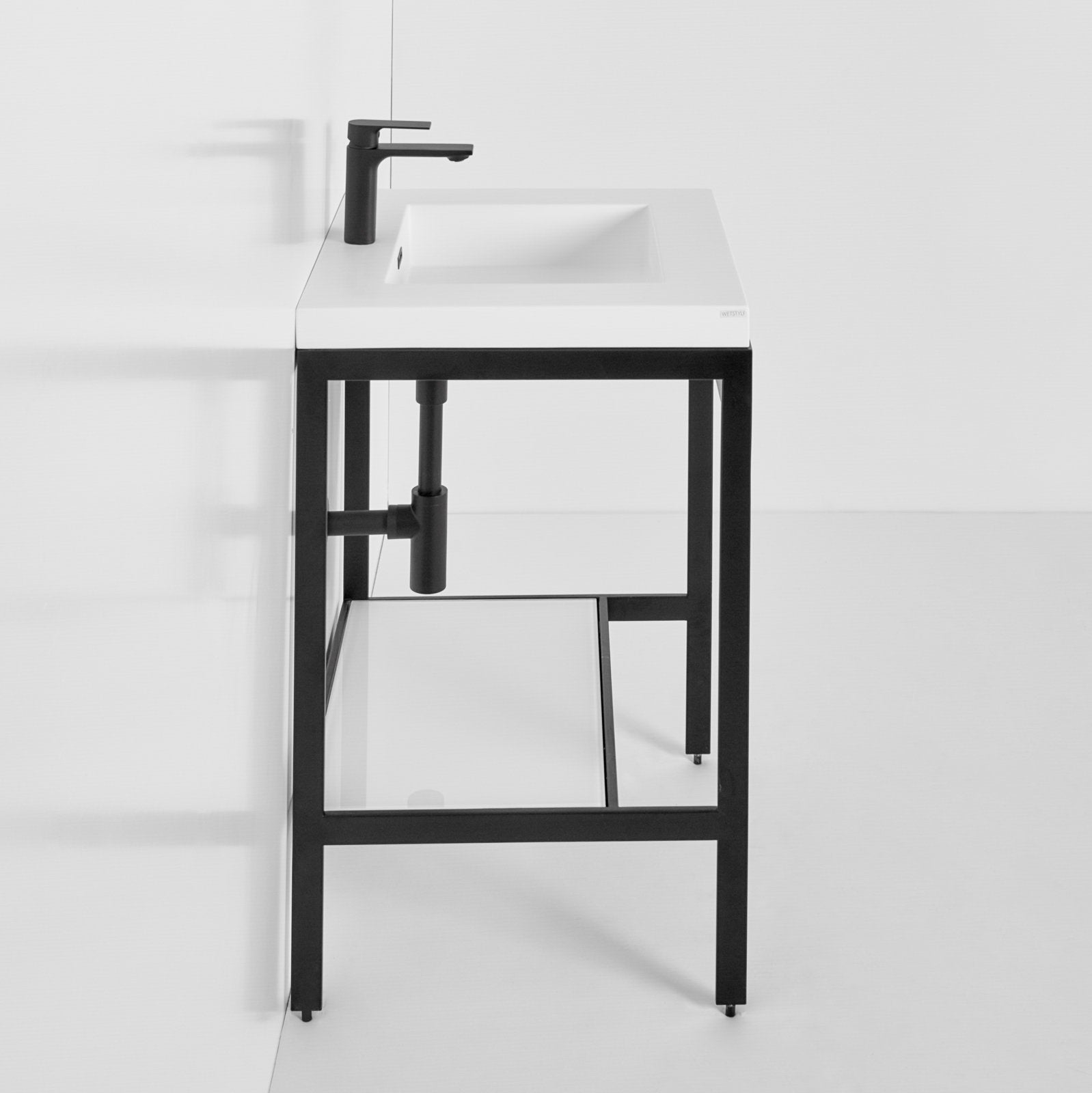 Freestanding modern single-sink bathroom vanity with a white stone rectangular integrated sink, matte black faucet, open black metal frame, exposed P-trap plumbing and a lower glass shelf.