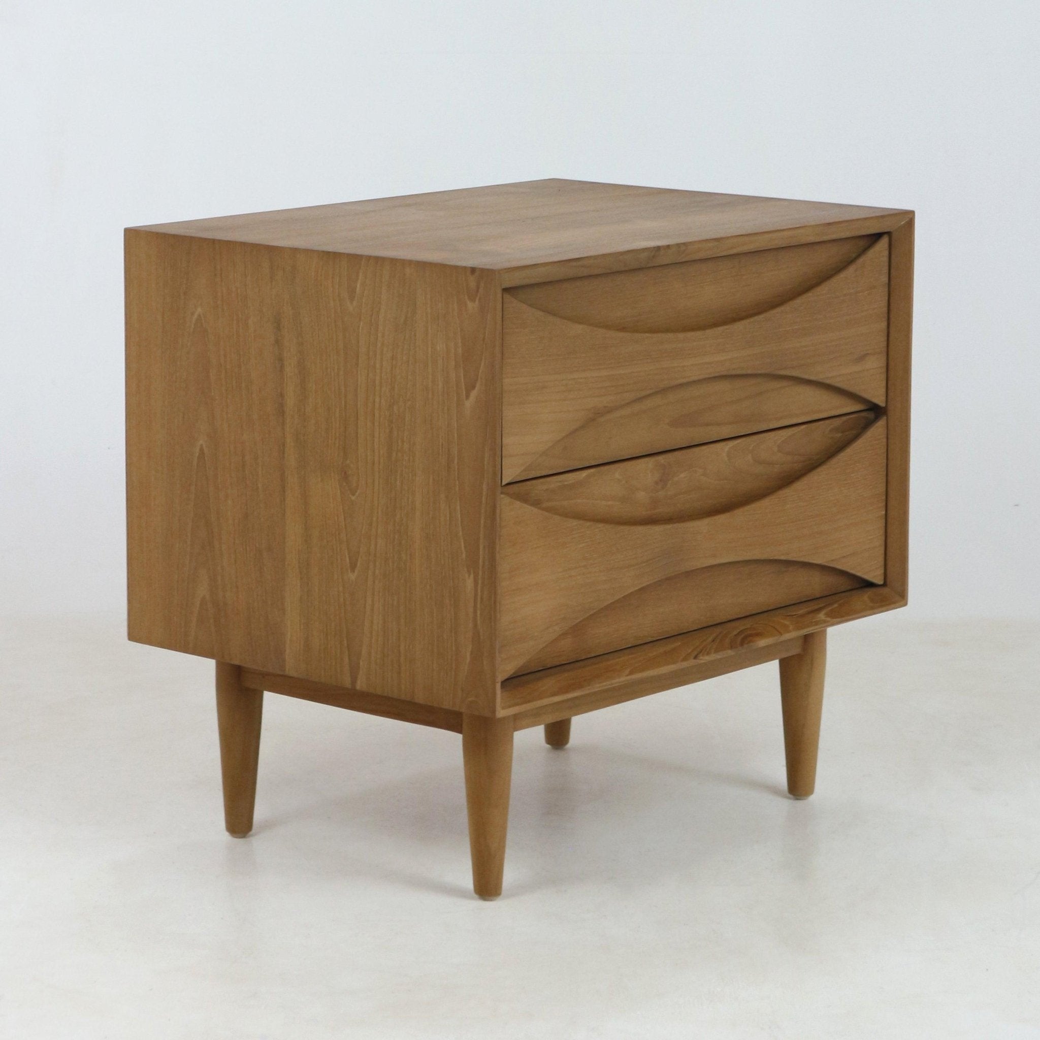 Solid teak wood mid-century modern nightstand in a natural finish with three drawers featuring carved recessed curved pulls and tapered legs, photographed on a white studio background.