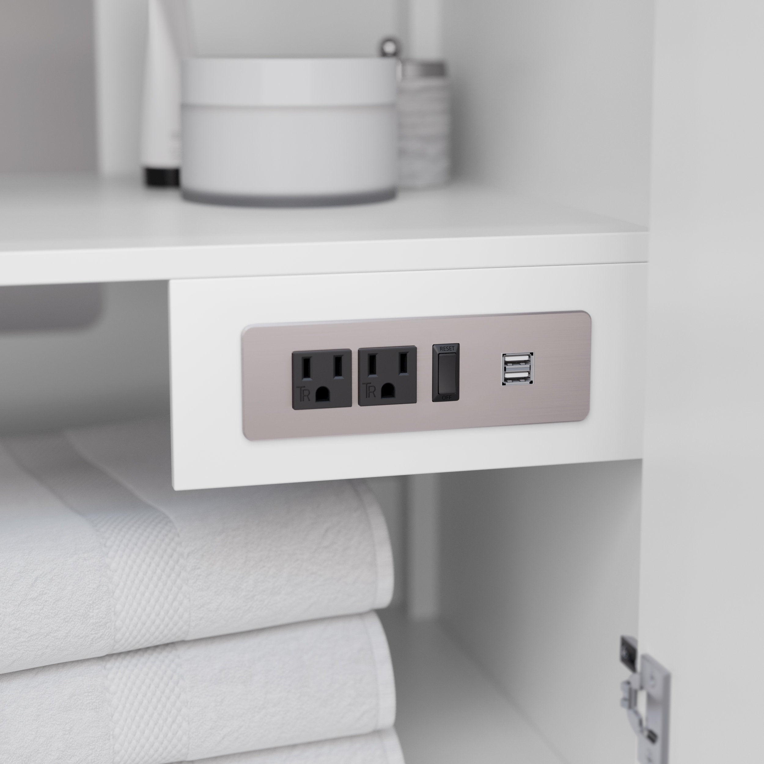 Close-up of a stone-colored power panel inside a white wooden bathroom vanity showing two black electrical outlets, a reset/GFCI switch and dual USB ports, with folded white towels below and toiletries above.