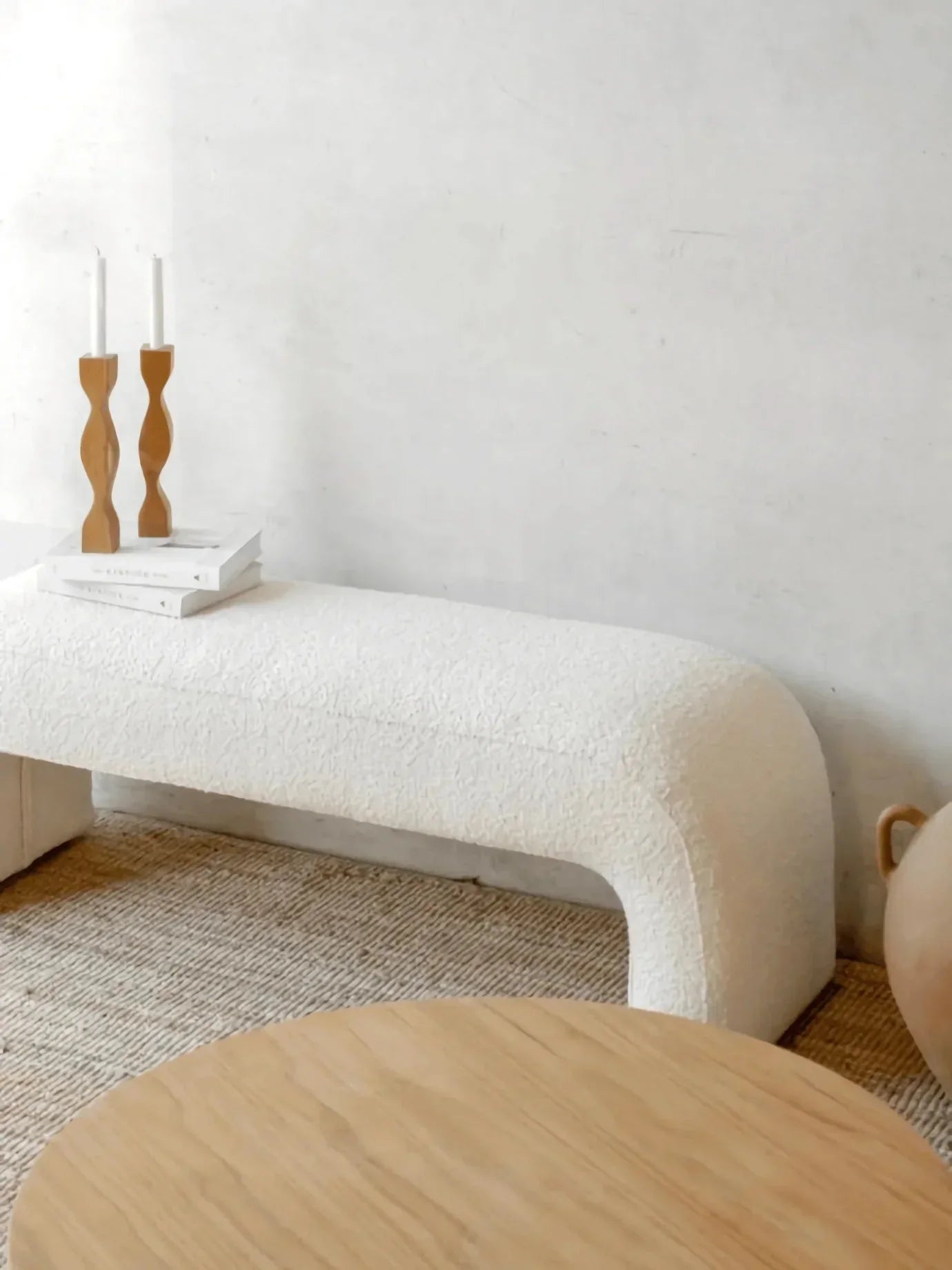 75-inch cream boucle upholstered bench with rounded arch legs on a natural woven rug, styled with stacked books, sculptural wooden candlesticks and a round light oak coffee table.