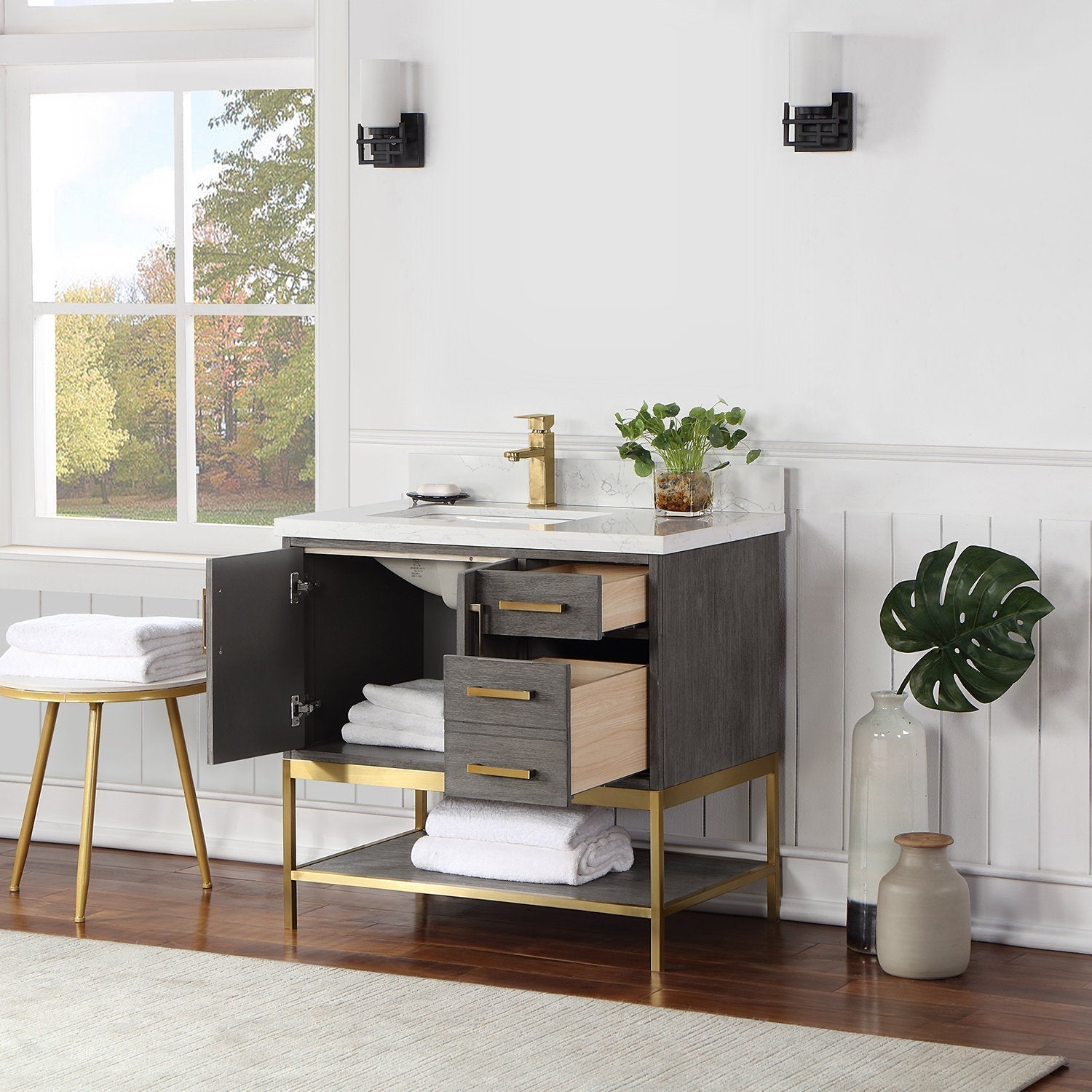 Modern solid wood bathroom vanity with stone top and single white ceramic sink, gray wood cabinet on a brass metal base with brass hardware and faucet; open door and drawers reveal neatly folded towels.