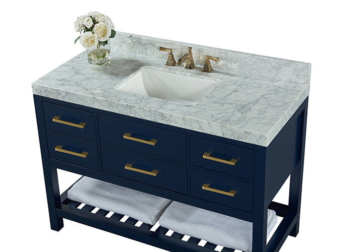 Navy solid-wood bathroom vanity with a gray marble stone top, recessed white ceramic sink, brushed gold faucet and handles, six front drawers and a slatted lower shelf holding folded towels and a small vase of white flowers.