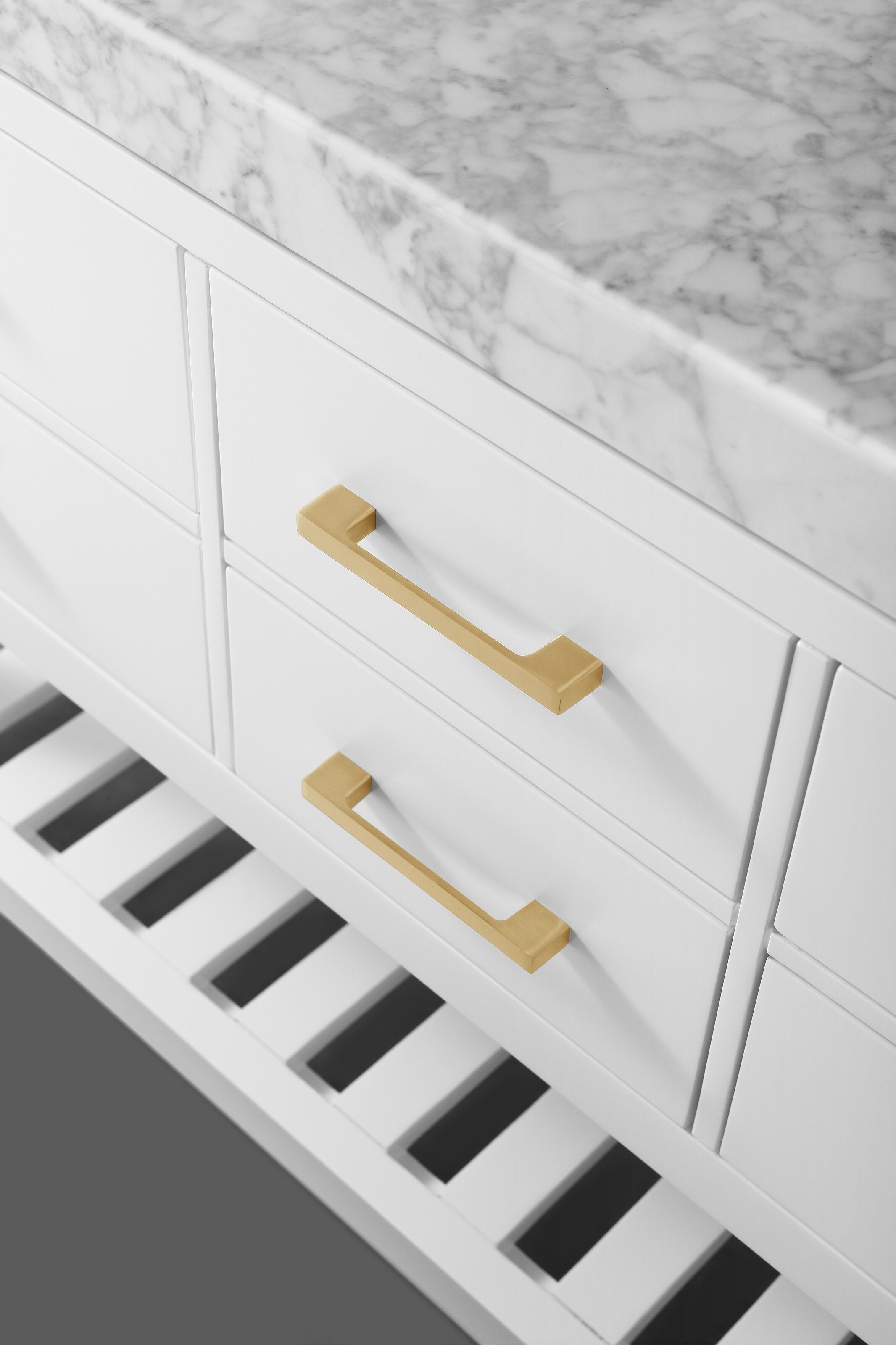 Close-up of a modern white solid wood bathroom vanity with a veined marble stone top, two gold drawer pulls and a slatted open shelf below.
