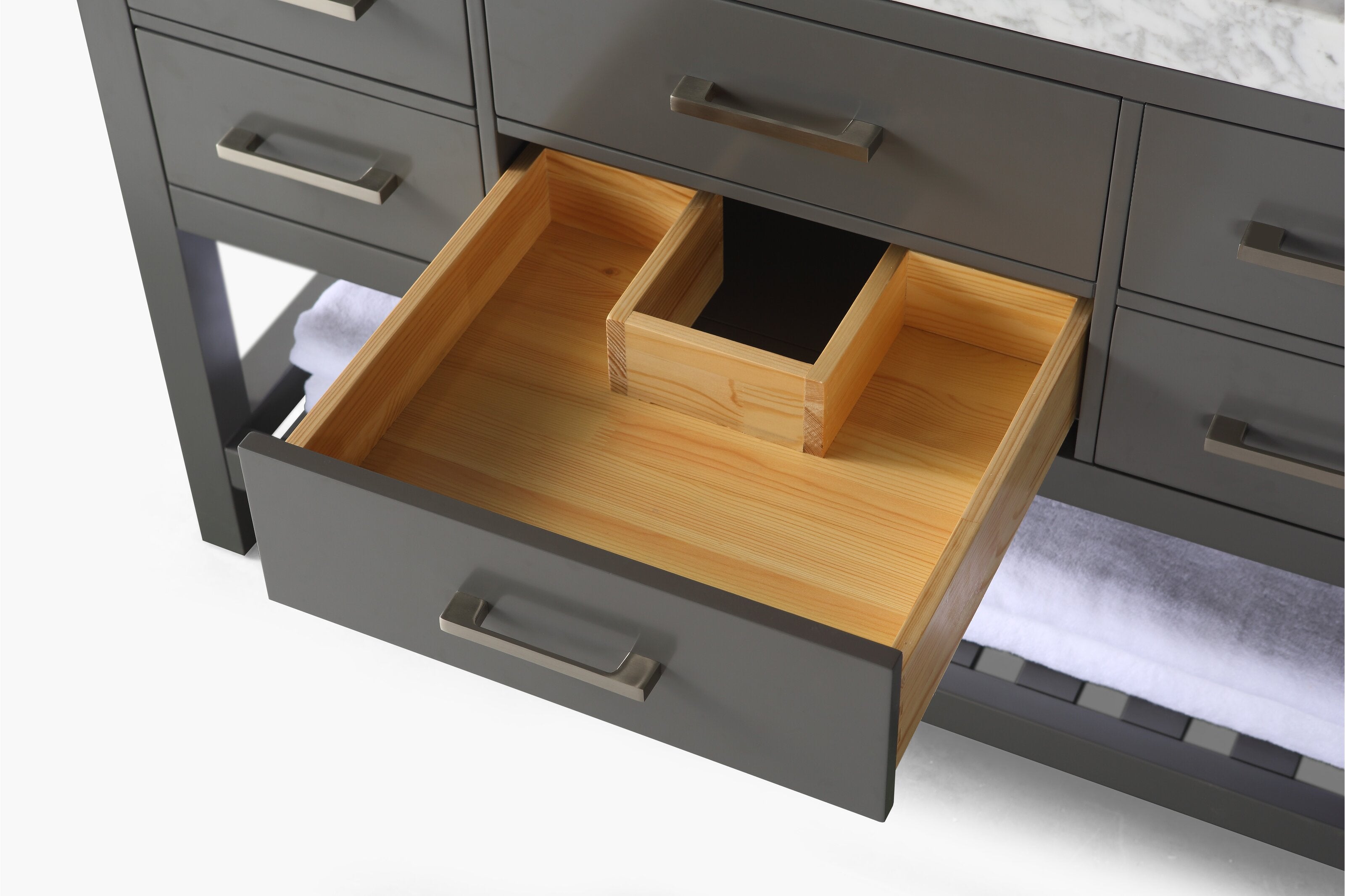 Close-up of a modern stone-colored bathroom vanity with an open drawer revealing a solid wood interior and center organizer, brushed metal handle, marble countertop edge and a lower shelf with a folded white towel.