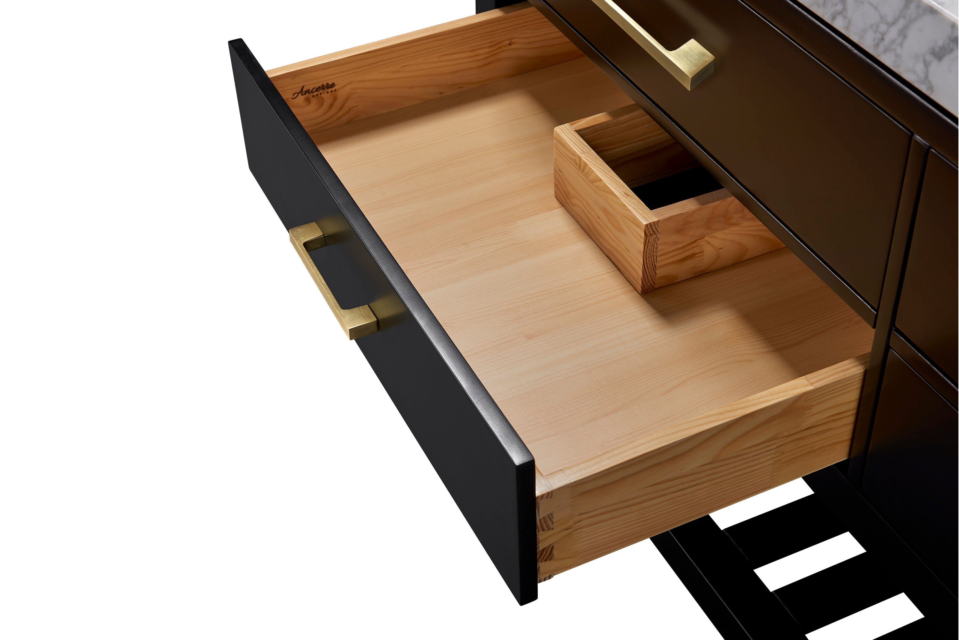 Open black bathroom vanity drawer revealing light solid wood interior with dovetail joints and a small wooden organizer, brushed gold metal handle on the matte black face and a glimpse of stone countertop and slatted shelf.