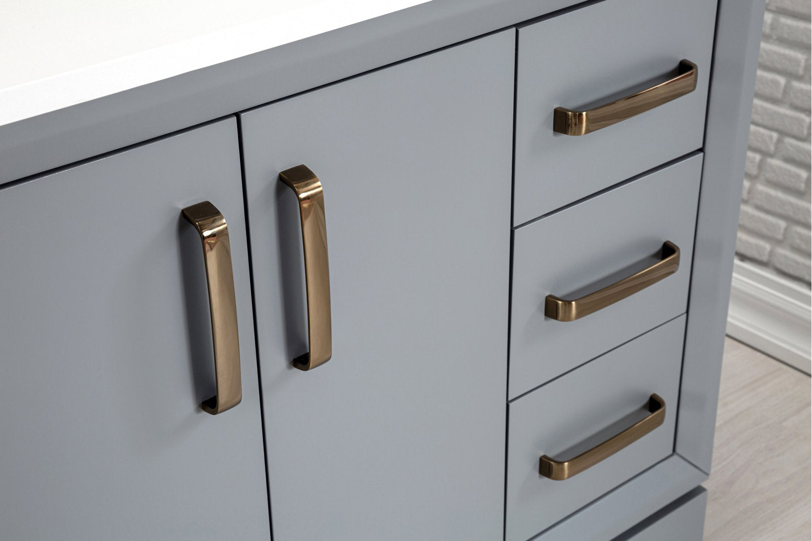 Close-up of a modern stone-colored solid wood bathroom vanity showing matte gray-blue cabinet doors and stacked drawers with brushed brass handles and a white stone countertop edge.