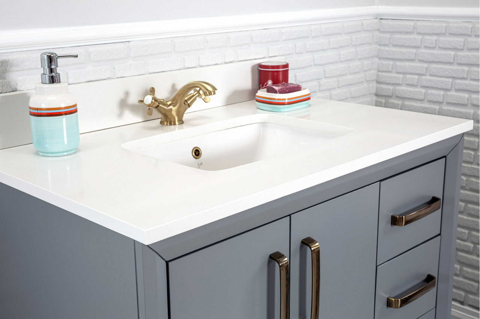 Close-up of a modern solid wood gray bathroom vanity with a white stone countertop and integrated rectangular sink, antique brass faucet and matching brass handles, with a textured white brick backsplash.