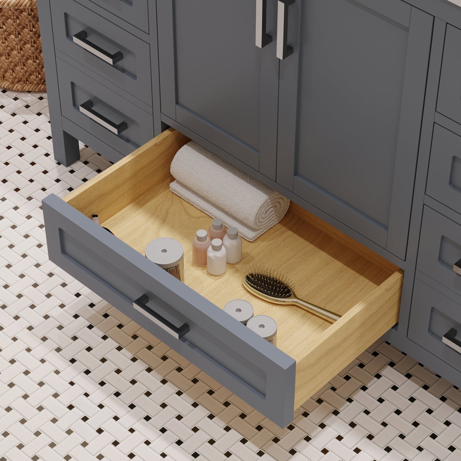 Open stone-colored modern bathroom vanity drawer revealing a natural solid wood interior with a rolled white towel, small toiletry bottles and gold-tone hairbrush; gray cabinet front and metal bar pulls over white mosaic floor.