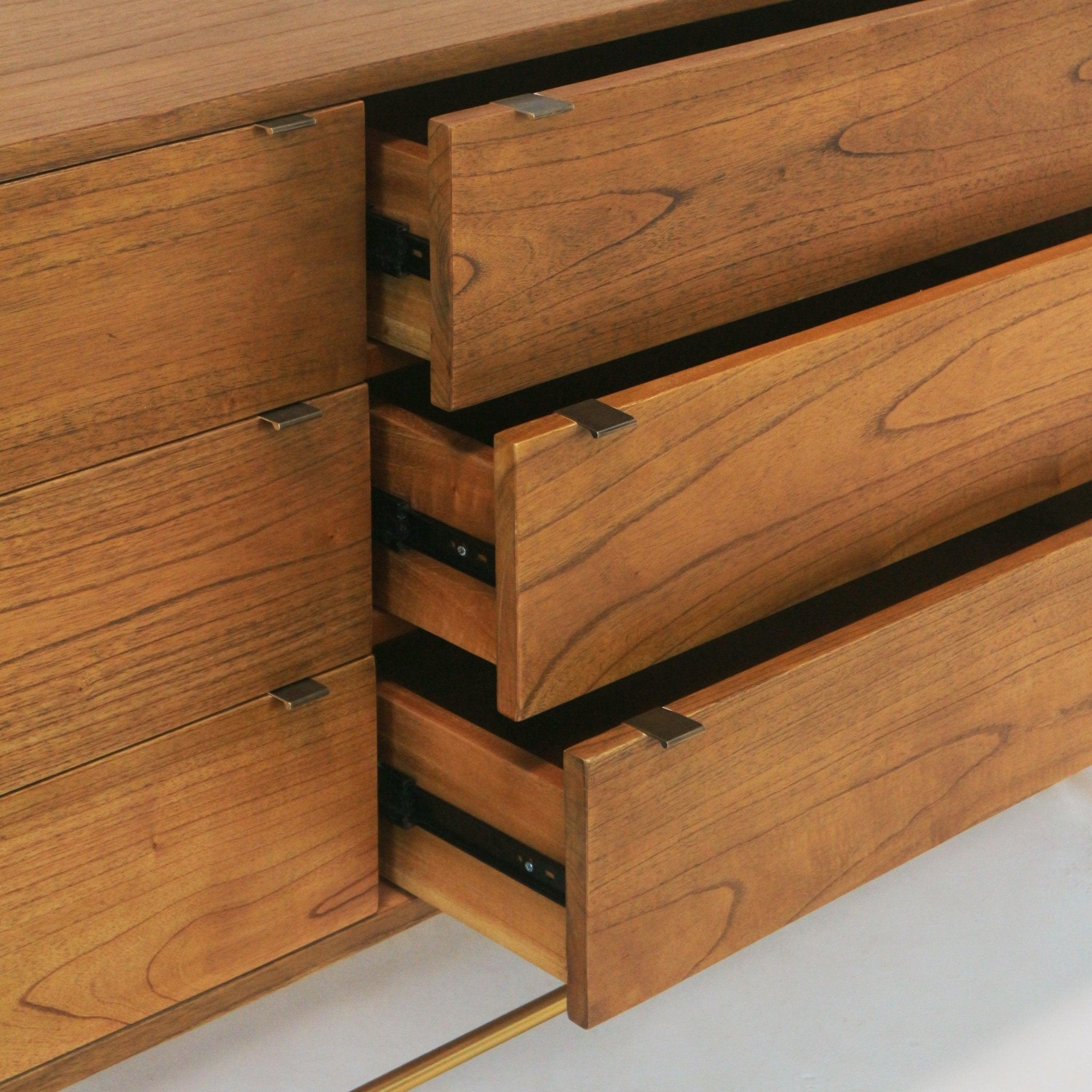 Close-up of three partially open natural teak drawers on a mid-century modern sideboard, showing warm wood grain, small brass rectangular pulls, visible metal drawer slides and a slim brass-toned bar base.