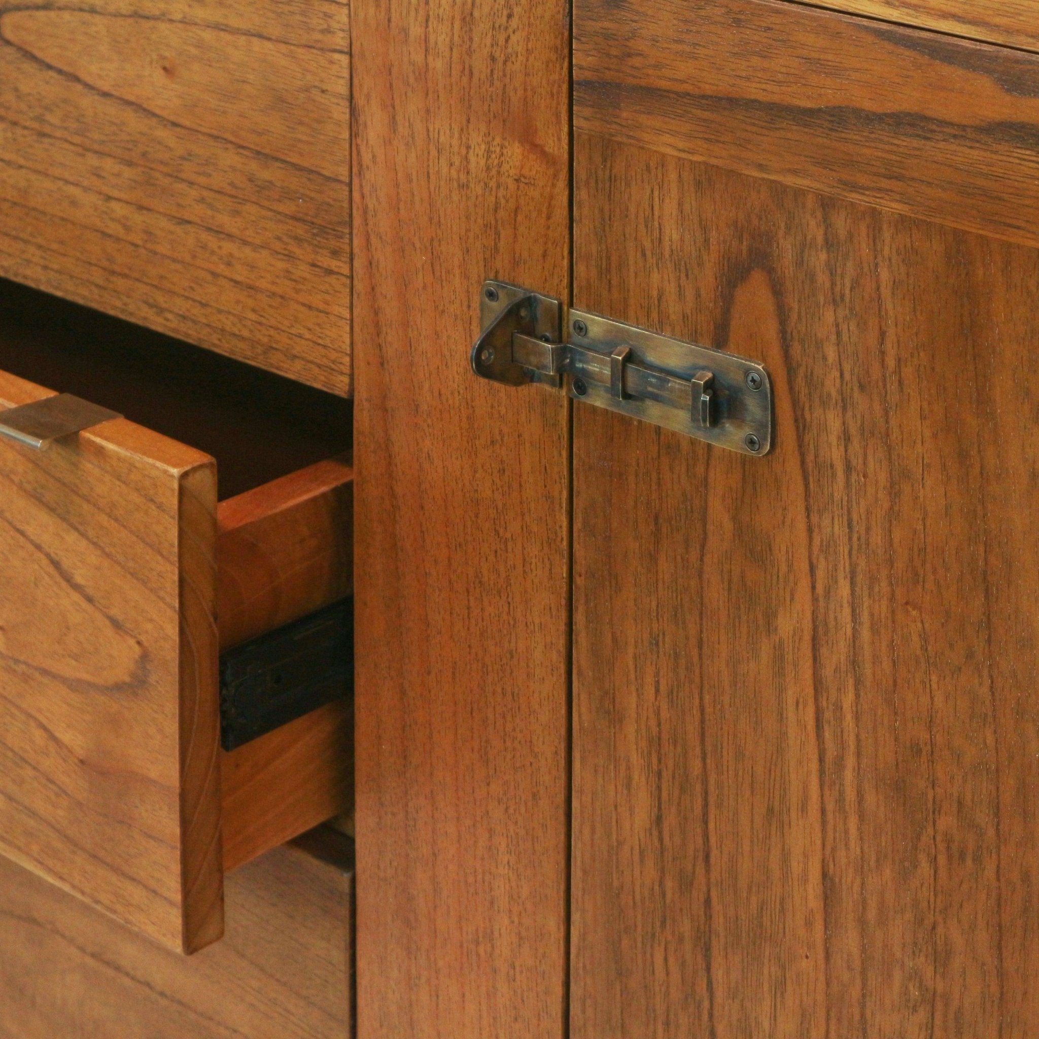 Close-up of a mid-century modern natural teak sideboard showing warm wood grain, a partially open drawer and an aged brass sliding latch on the cabinet door.