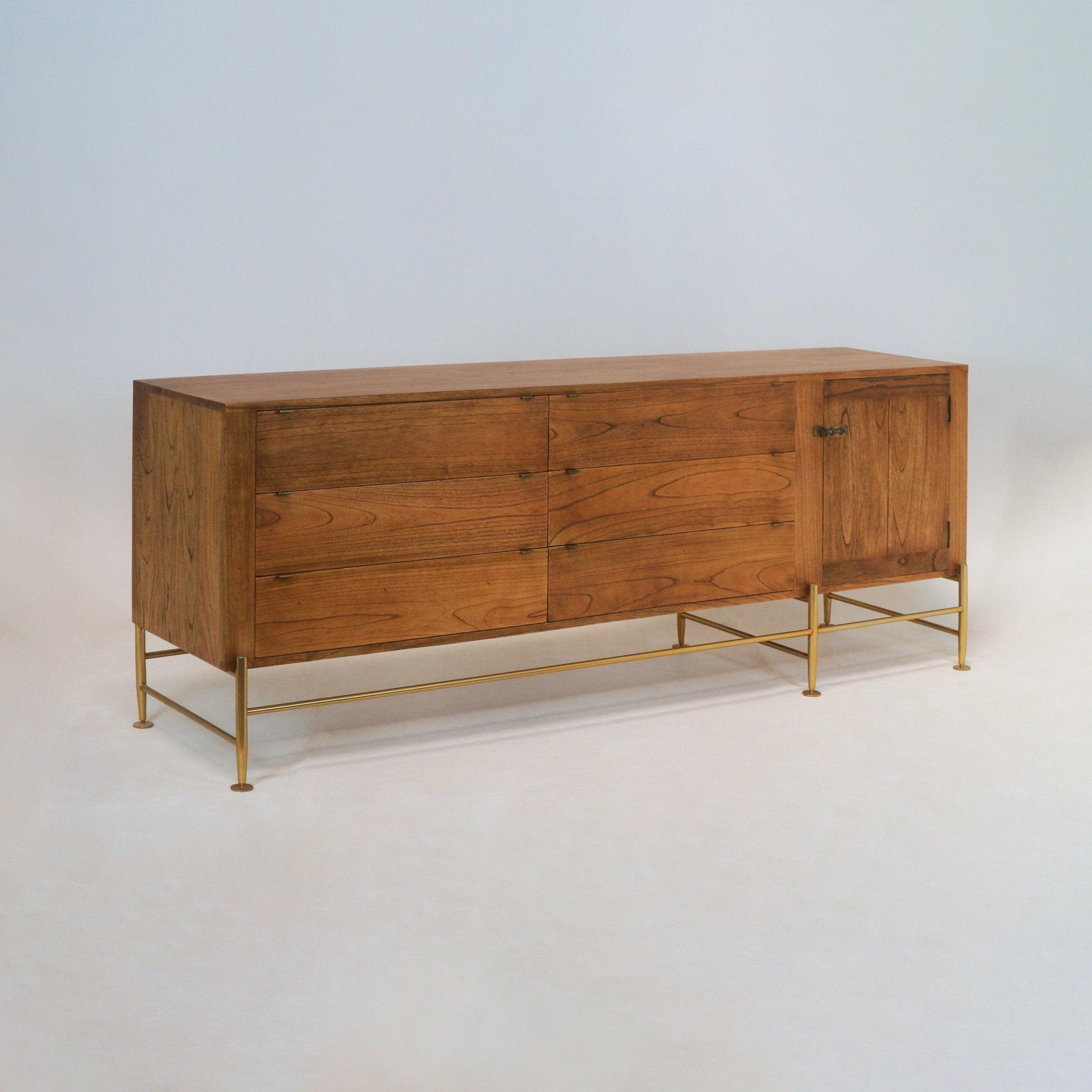 Mid-century modern teak sideboard with six wide horizontal drawers and a single right-side cabinet door, warm natural teak grain, small recessed metal pulls, and a slim brass leg frame with crossbar supports.