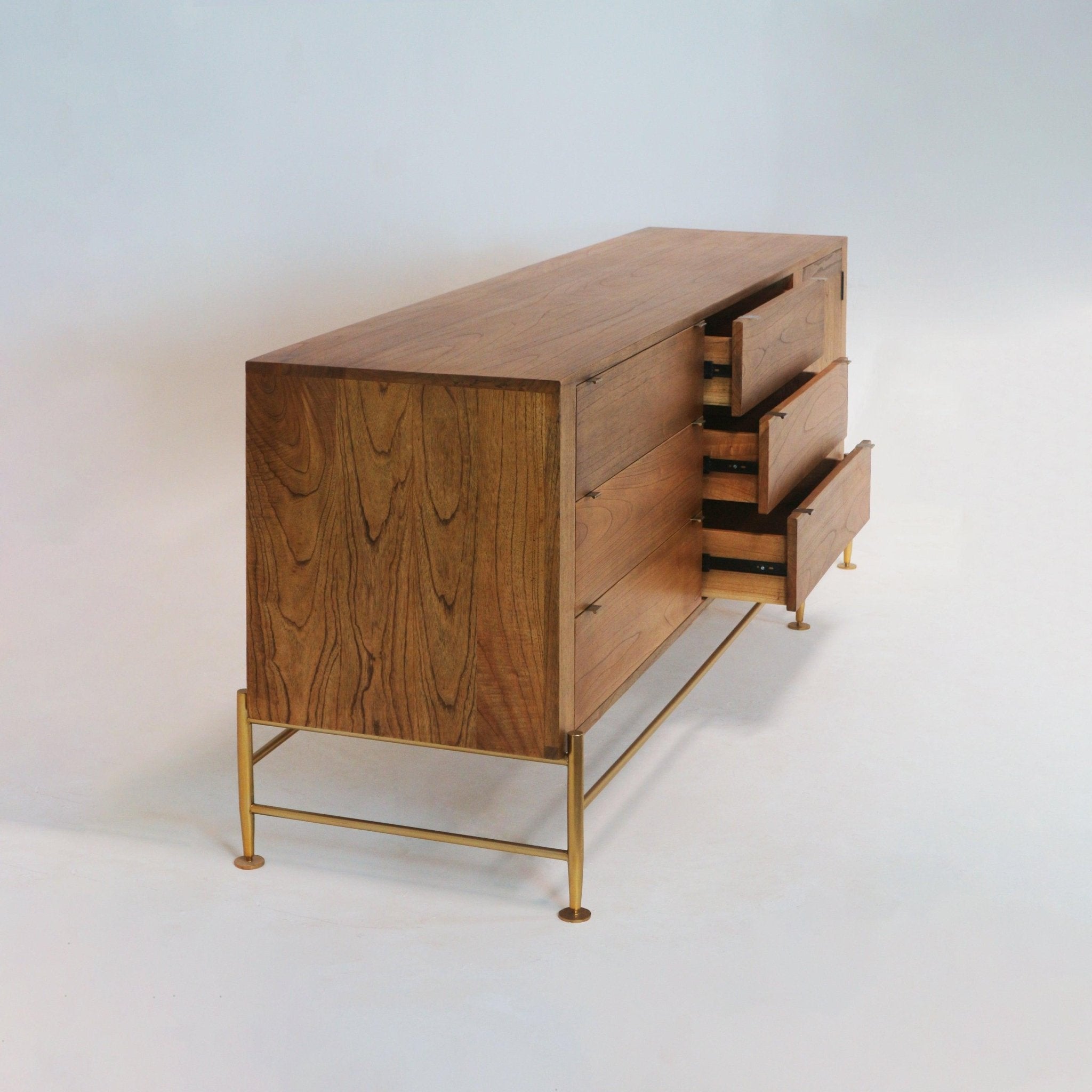 Three-quarter view of a long natural teak sideboard with three partially open drawers, visible wood grain and a slim brass metal base and legs in a mid-century modern silhouette on a white studio background.