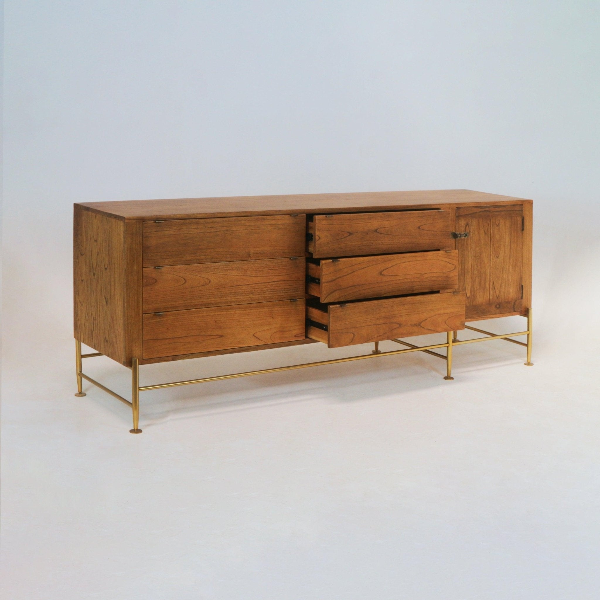 Natural teak midcentury sideboard showing warm wood grain, three center drawers partially open, additional left drawers and a right cabinet door, mounted on slim brass metal legs with a horizontal stretcher.
