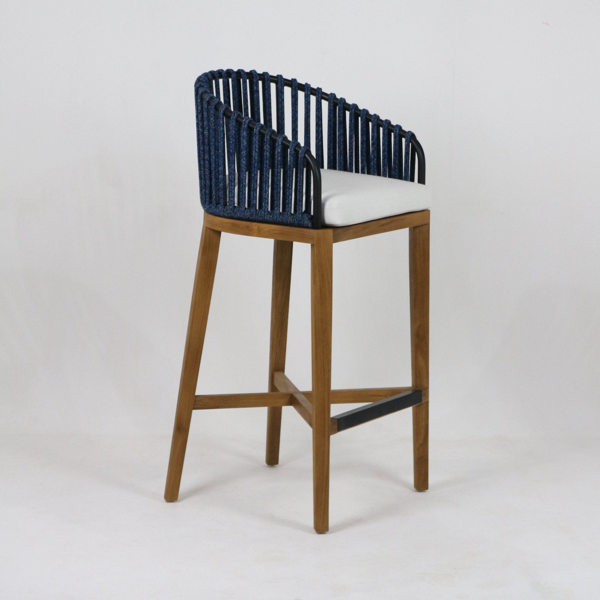 Outdoor teak bar stool with a navy woven rope back, white seat cushion, tapered teak legs and a black metal footrest, photographed on a plain studio background.
