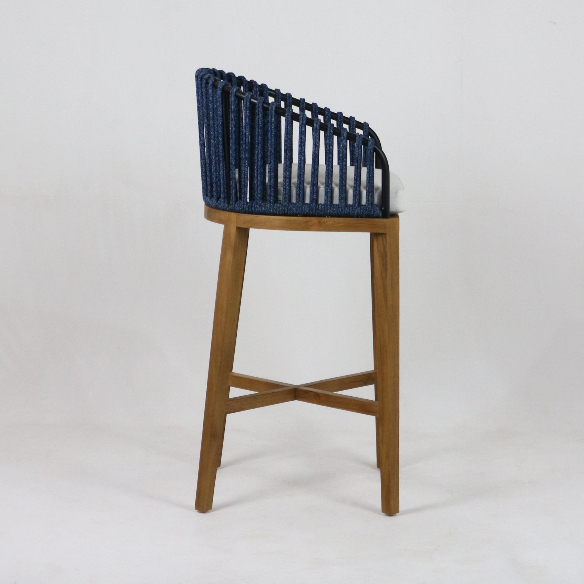Side view of a teak outdoor bar stool with a navy blue woven rope back, black metal support, white seat cushion and angled teak legs with an X-shaped cross stretcher.