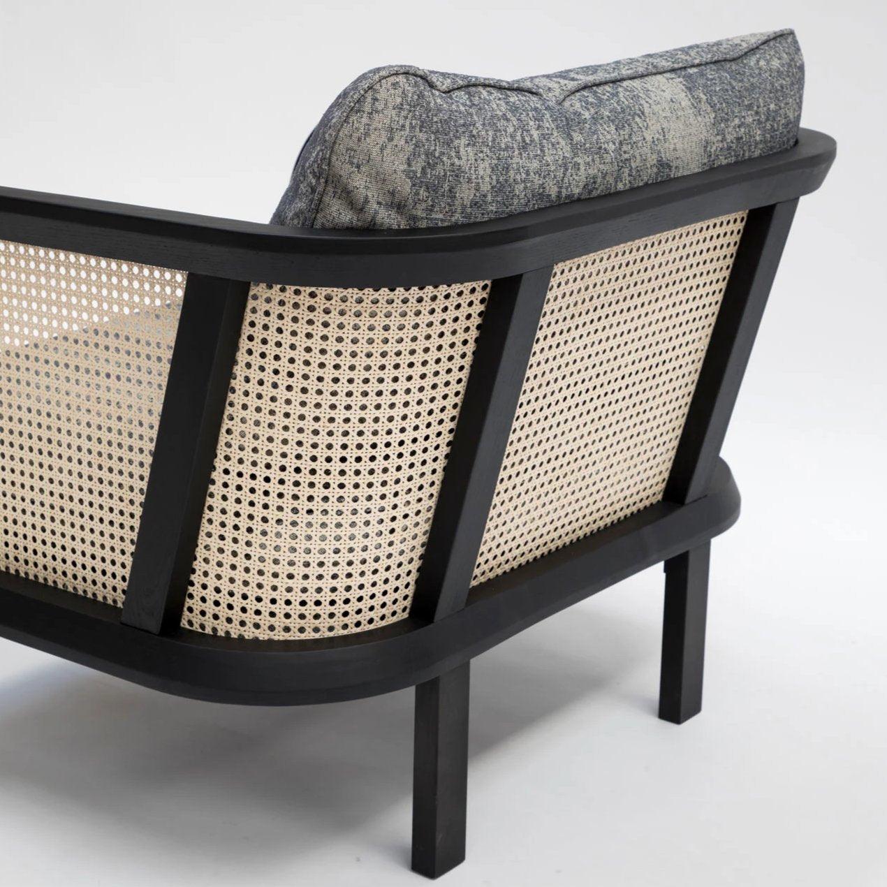 Close-up of the modern chair's rear showing a black wooden frame, natural woven cane back panels and a textured gray-blue fabric cushion resting on straight black legs.