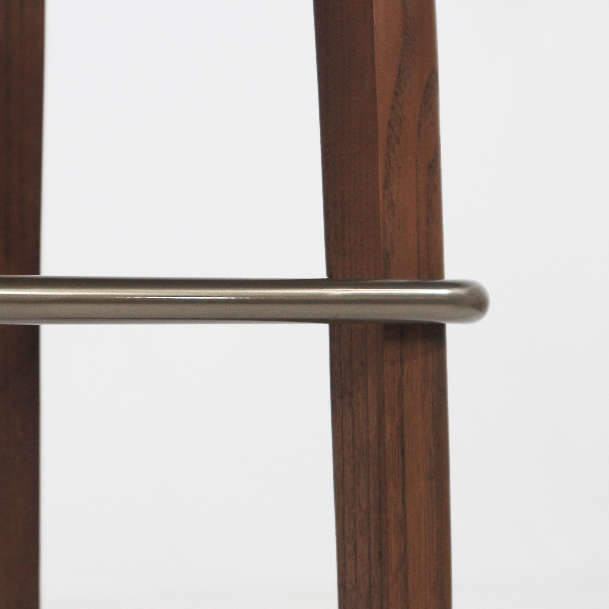 Close-up of a brushed metal footrest bar with a rounded end mounted between two walnut-stained wooden stool legs against a white background.