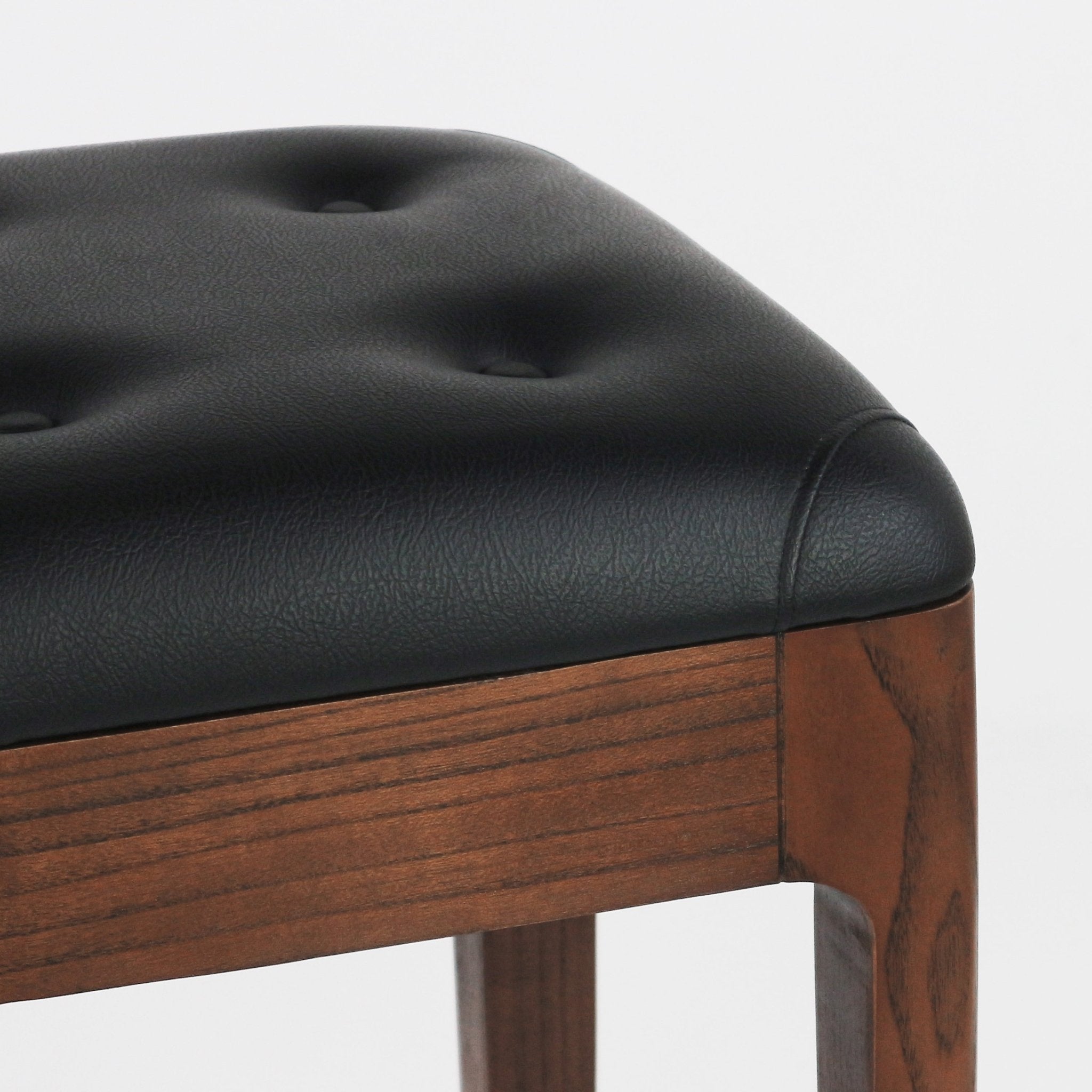 Close-up of a tufted black leather seat cushion with button detailing atop a walnut-finished wooden bar stool frame and square leg.