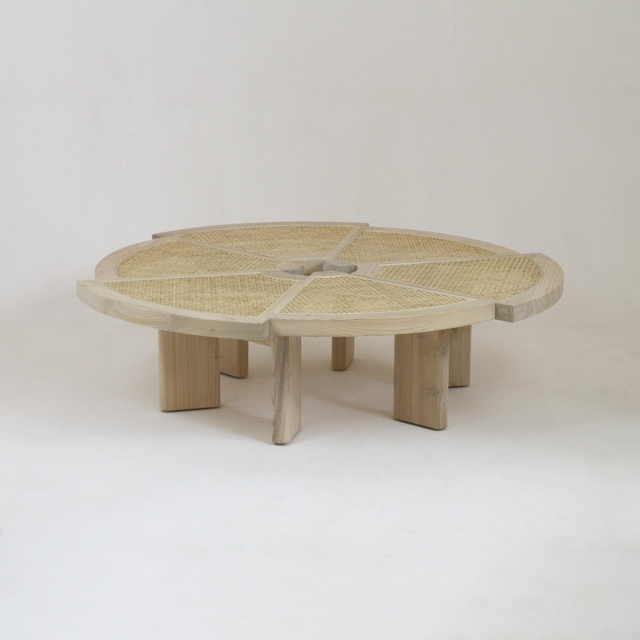 Low round coffee table in natural oak with woven rattan (cane) inset top divided into six pie-shaped panels and chunky sculpted legs, photographed on a neutral studio background.