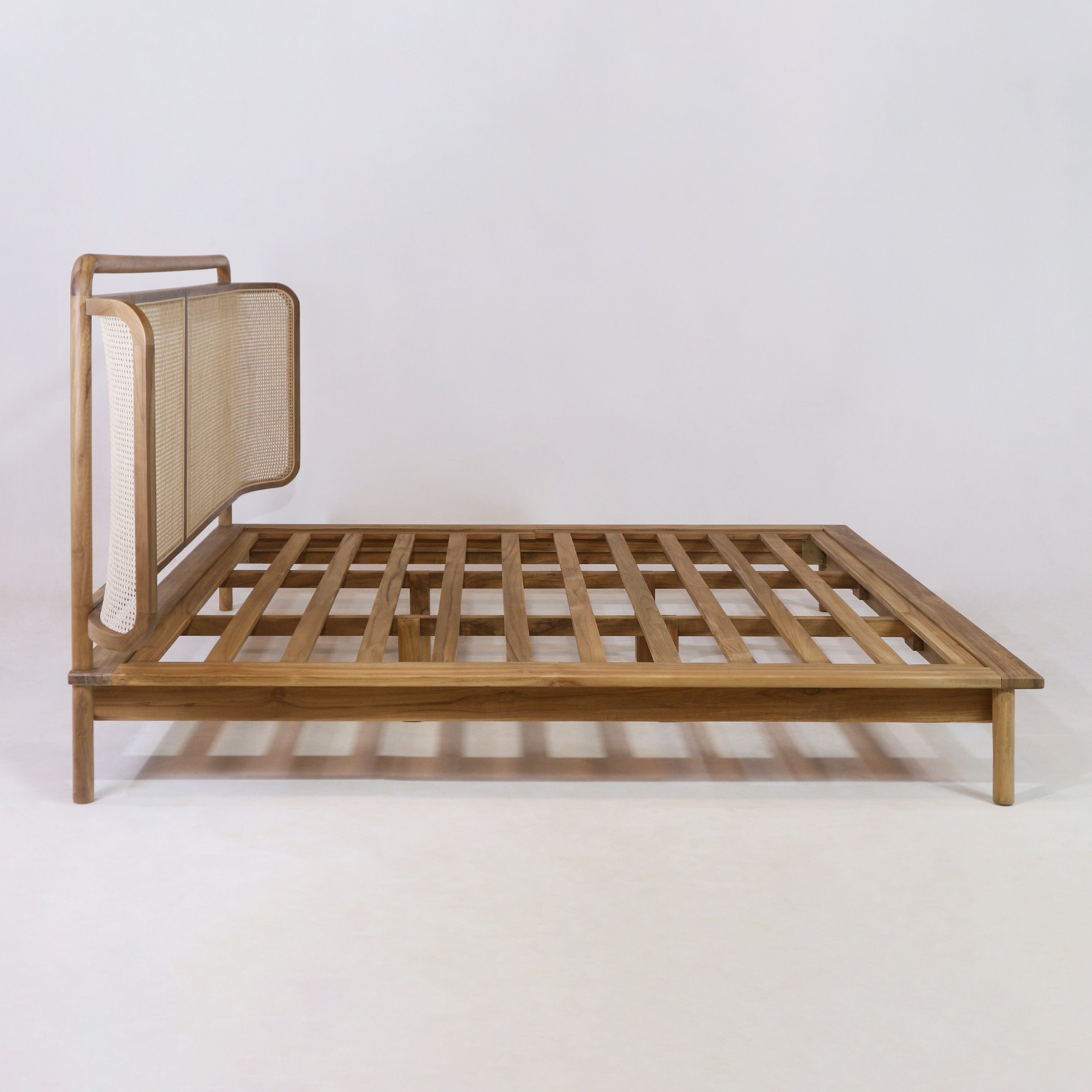 Low-profile teak platform bed frame with a woven rattan headboard, open slatted teak base and rounded wooden frame in a natural teak finish, photographed on a white studio background.