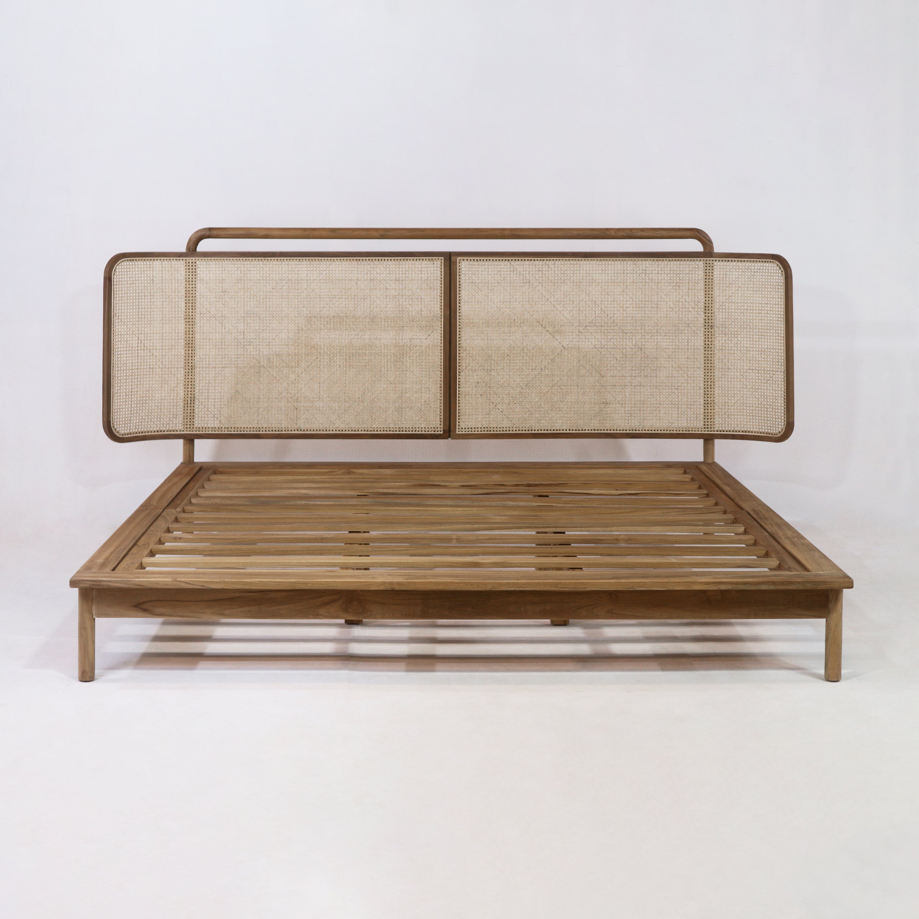 Front view of a low-profile teak platform bed with slatted wooden base and woven rattan cane headboard panels in a natural teak finish, presenting a boho-style look against a white background.