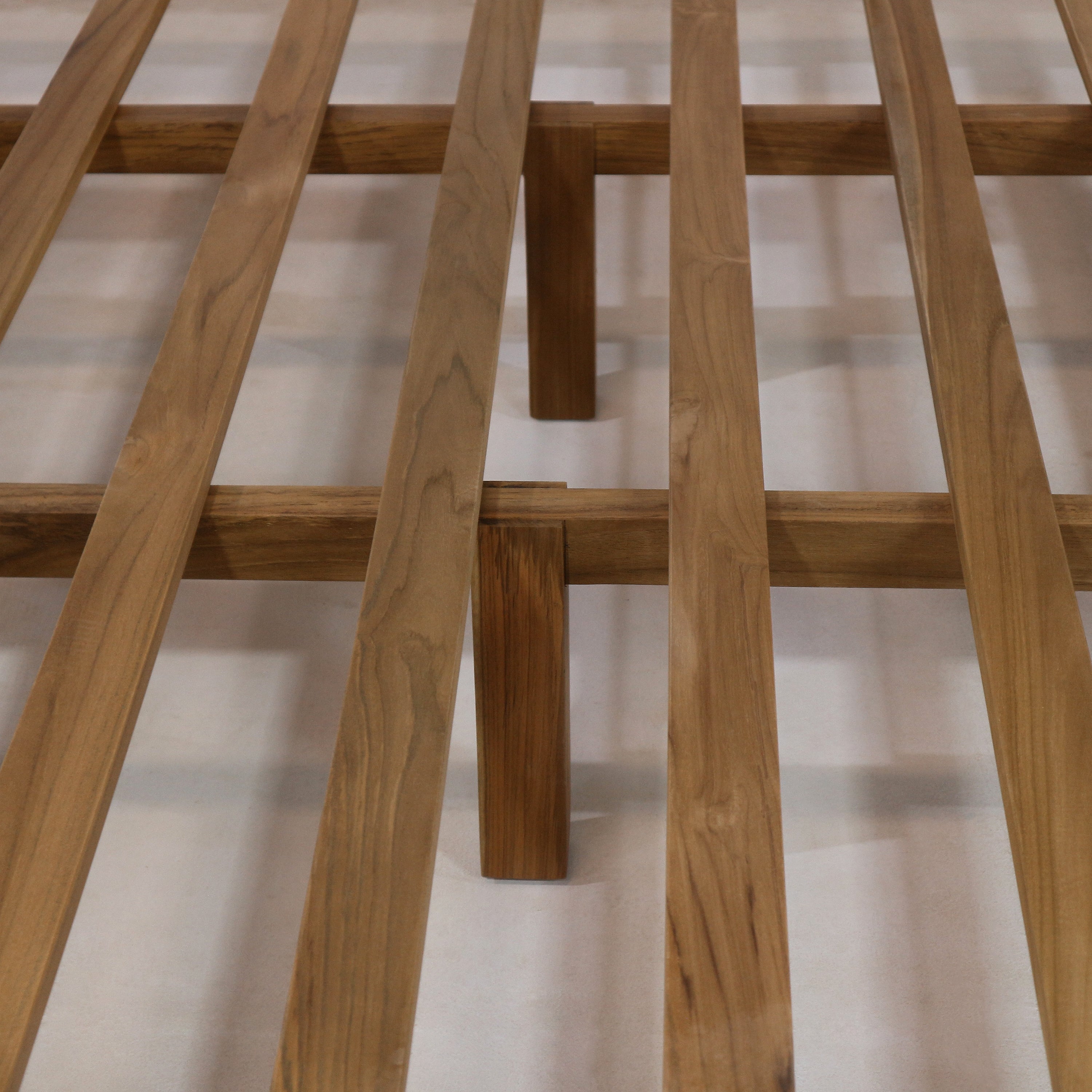 Close-up of a teak slatted platform bed frame showing evenly spaced wooden slats and a central support leg in a natural teak finish.