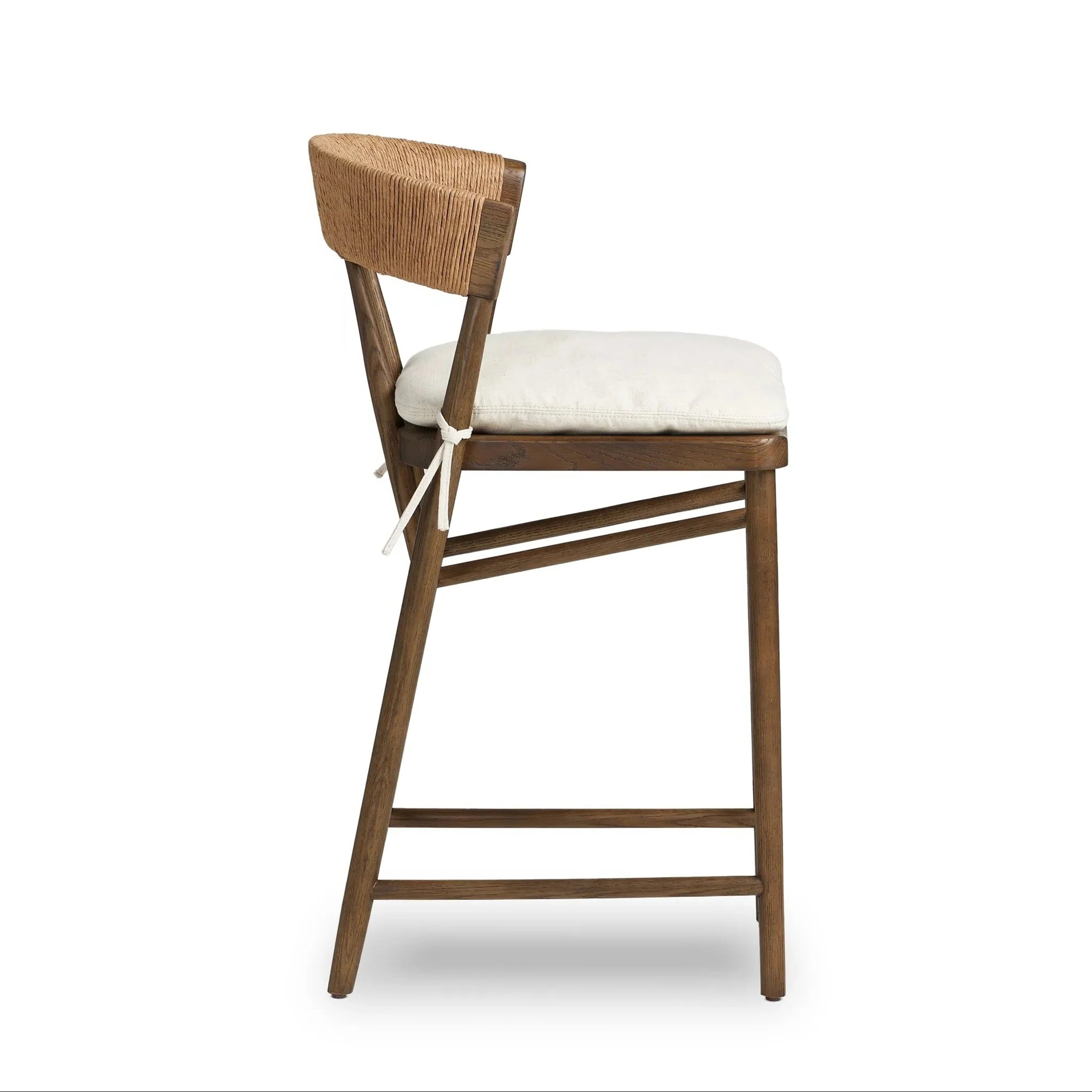 Side view of a natural wood counter-height stool with a cream cushioned seat tied with fabric ties and a rope-wrapped C-shaped backrest, angled legs and double footrest in a rustic farmhouse finish.