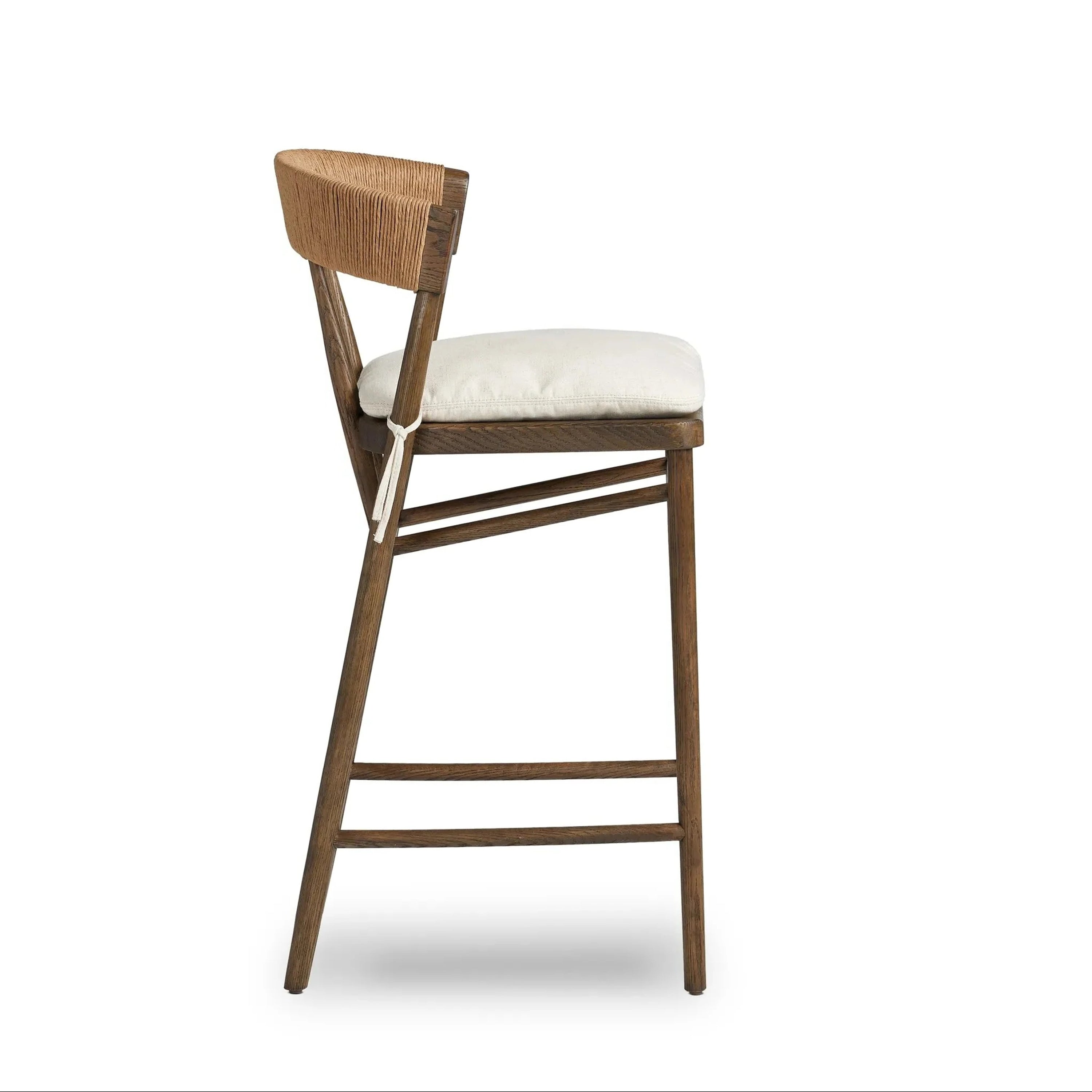 Side view of a natural wood counter stool with a woven rope C-shaped back, off-white cushioned fabric seat tied at the side, angled legs and double-step footrest bars in a rustic farmhouse finish.