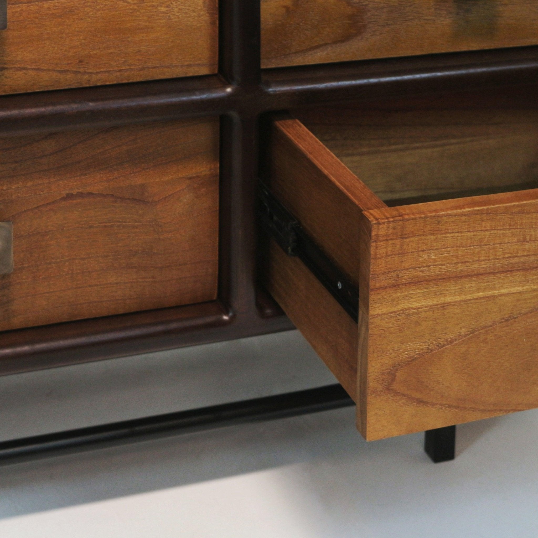 Close-up of an open teak drawer showing warm teak wood grain, a black metal drawer slide and dark iron cross-frame and leg of a mid-century modern sideboard.