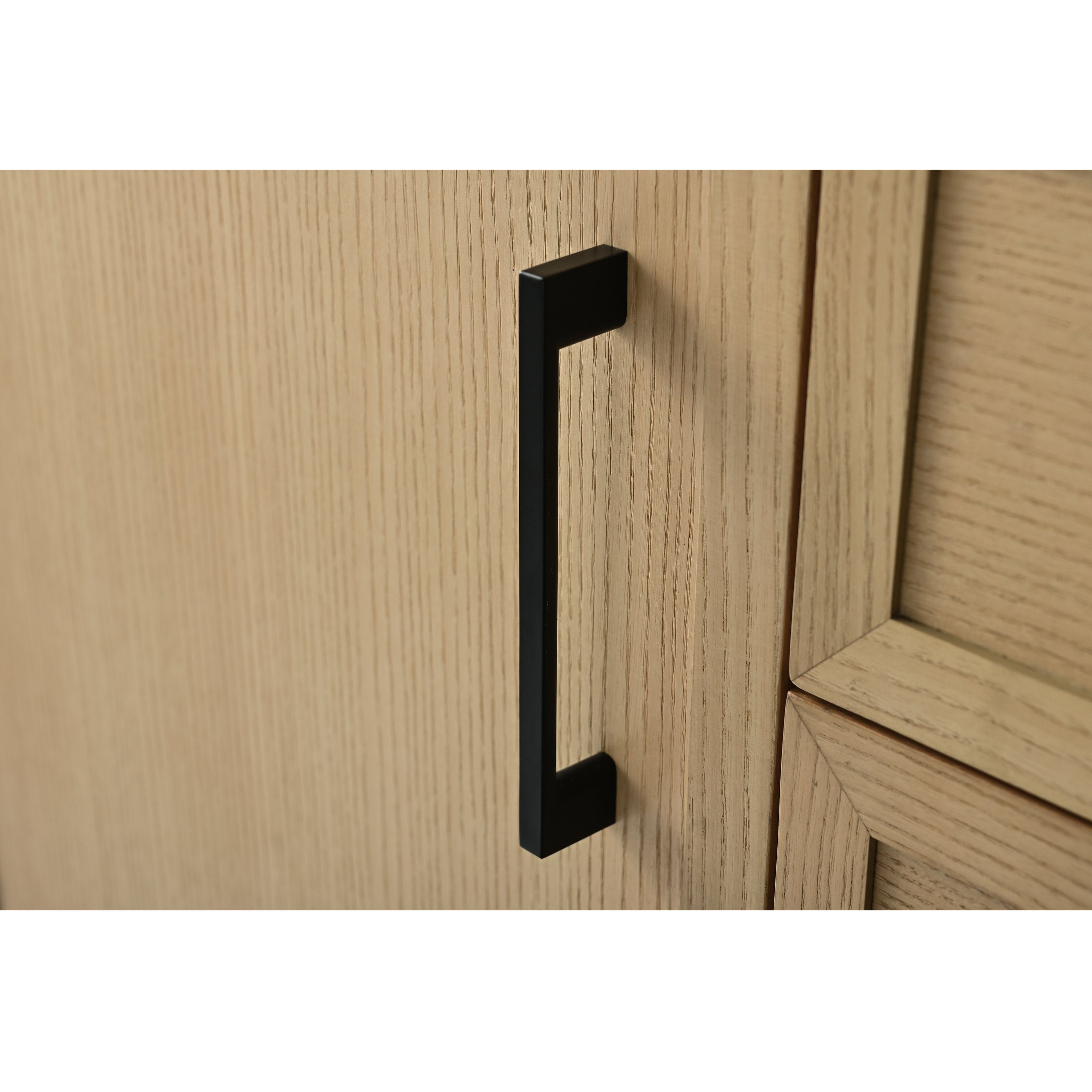 Matte black rectangular cabinet pull mounted vertically on a light stone-colored solid wood vanity door, showing wood grain texture and the adjacent recessed panel edge.
