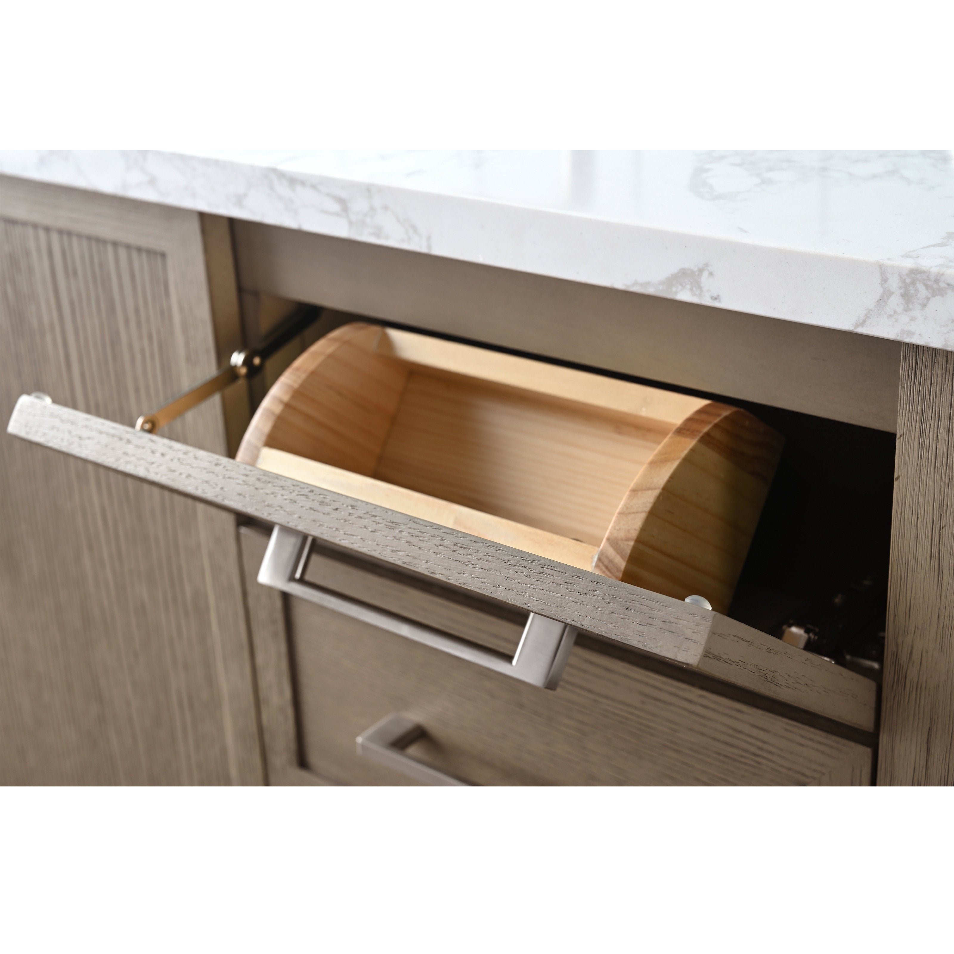 Close-up of a single-sink bathroom vanity tilt-out drawer with a marble-look stone countertop, stone-colored solid wood cabinet, brushed metal bar handle and a light natural wood internal bin.