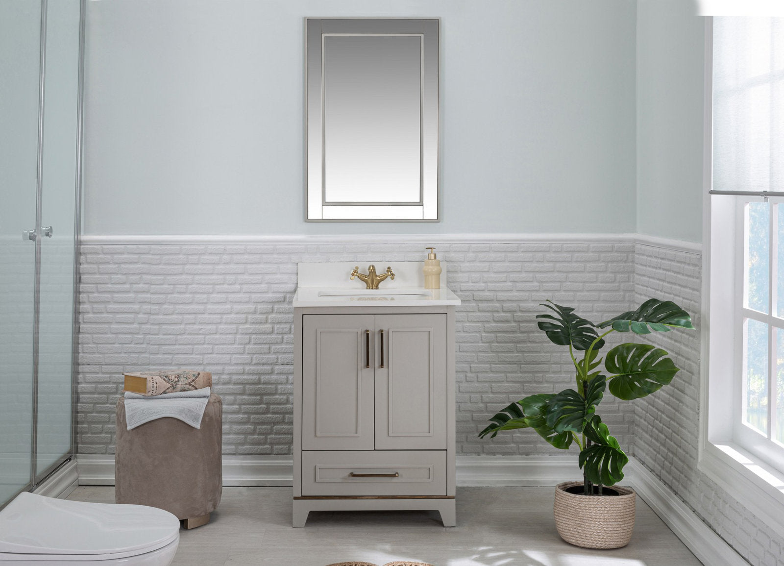 Freestanding modern solid wood bathroom vanity in a stone finish with white stone countertop, brass faucet and single sink; rectangular mirror above, potted plant and fabric ottoman in a light neutral bathroom.