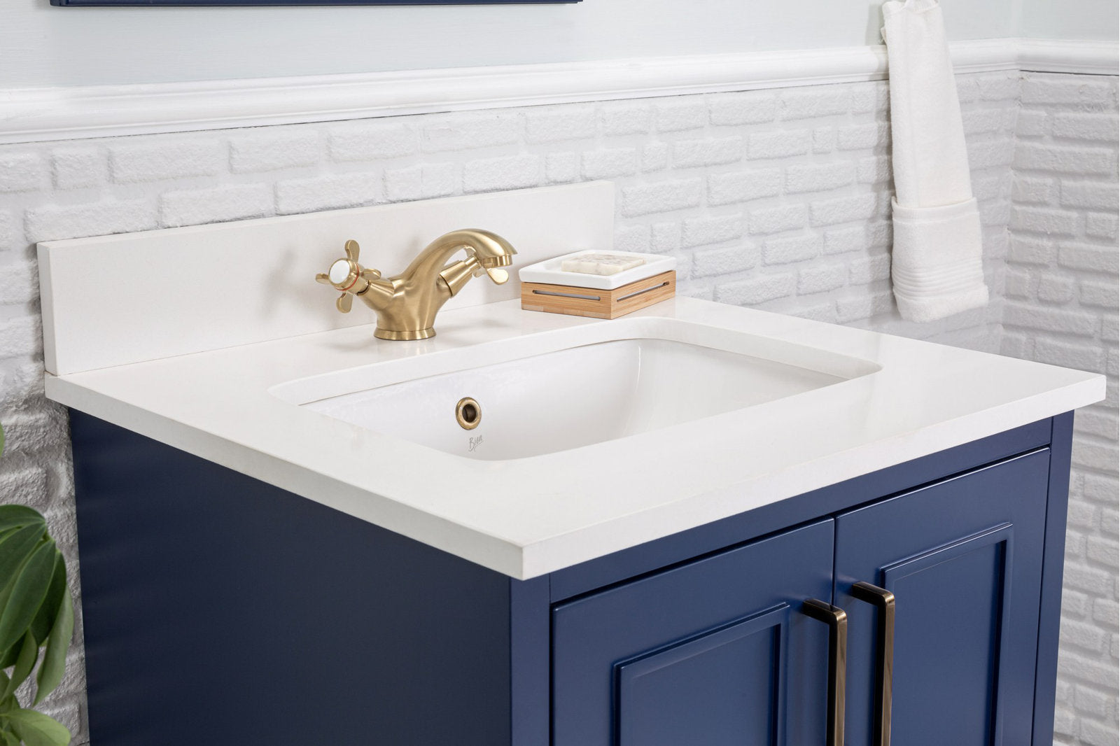 Close-up of a modern solid wood single-sink bathroom vanity with a stone top and integrated ceramic sink, navy blue cabinet, brass cross-handle faucet and matching brass pulls against a white textured brick backsplash.