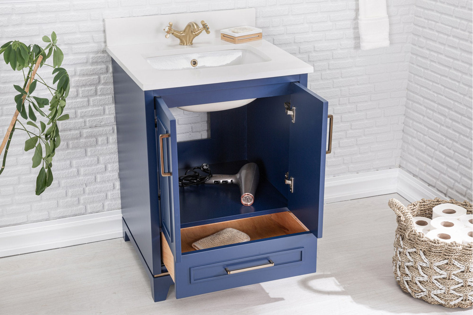 Modern navy solid-wood bathroom vanity with white stone countertop and integrated ceramic sink, brass faucet, open cabinet revealing a hair dryer and open drawer, shown against a white brick wall with woven toilet-paper basket.