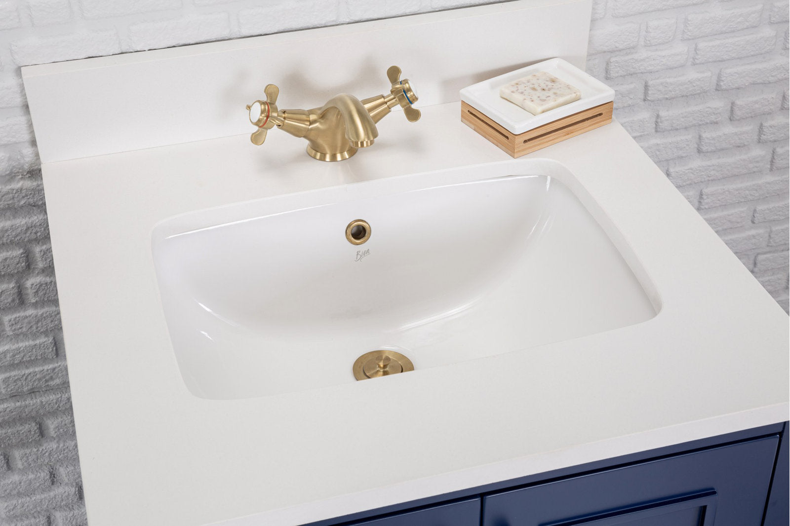 Top view of a modern single-sink bathroom vanity with a white stone countertop and inset ceramic sink, brass two-handle faucet and drain, wooden soap dish with soap, and navy-blue vanity drawers against a white brick backsplash.