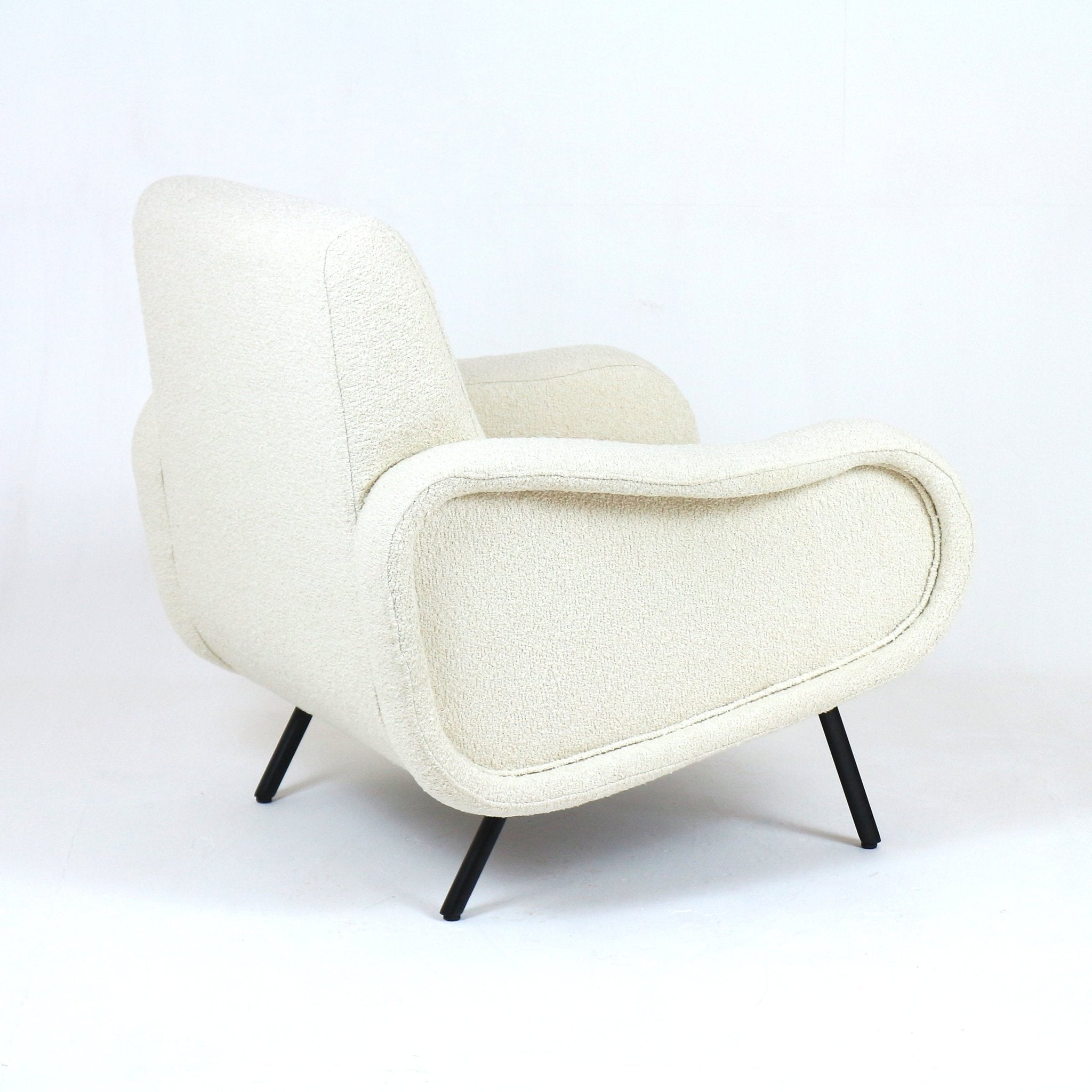 Rear three-quarter view of a modern white boucle upholstered arm chair with a rounded low back, curved padded arms and tapered black metal legs on a white studio background.