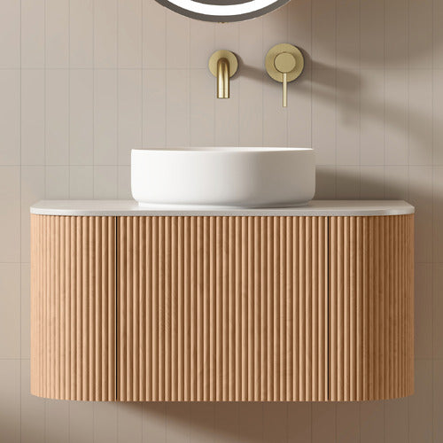 Floating fluted teak vanity with a rounded white ceramic vessel sink on a pale stone top, paired with wall-mounted brushed brass faucet against a neutral beige tiled wall.