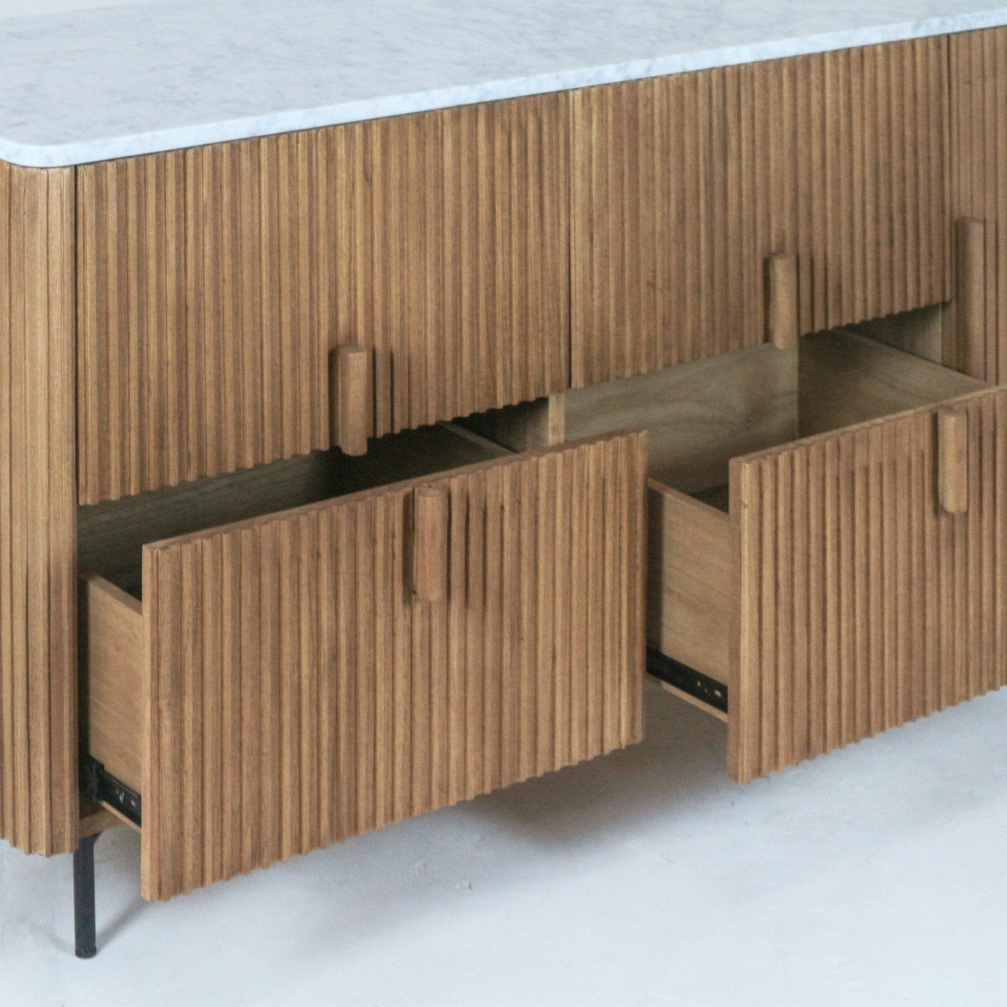 Close-up of a mid-century modern teak sideboard showing fluted vertical slat doors, two partially open drawers with round wooden pulls, a rounded white stone top, and slender black metal legs.