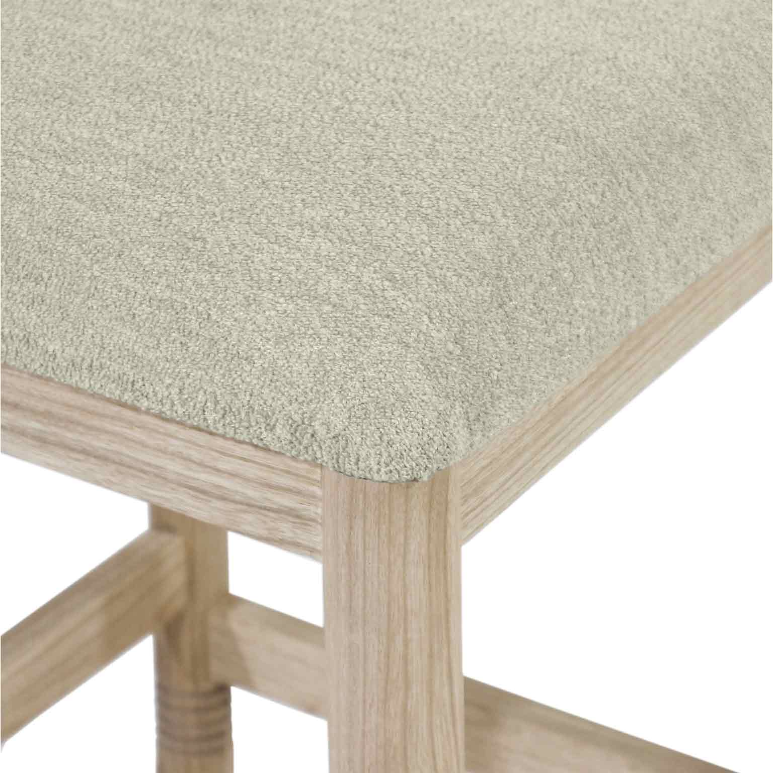 Close-up of a beige bouclé upholstered seat corner on a natural oak stool frame, showing textured boucle fabric, a rounded seat edge and the oak leg with integrated footrest.