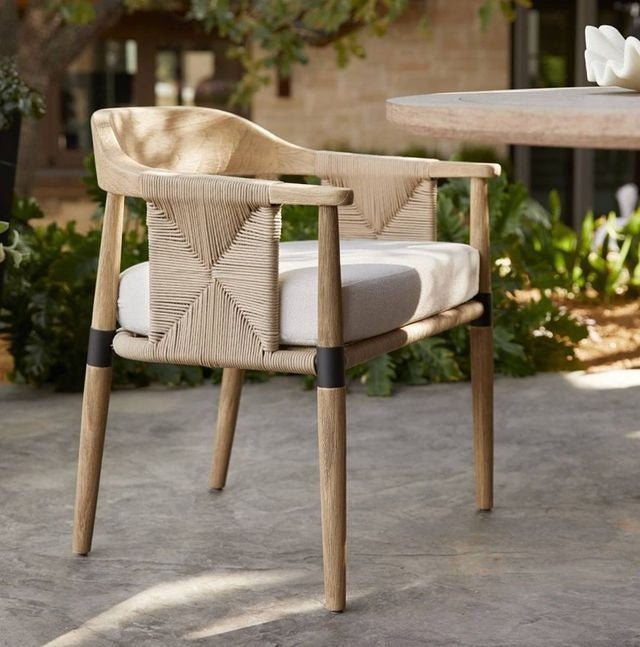 Natural oak dining chair with woven rope back and integrated arms, light beige performance-fabric seat cushion, tapered oak legs with black metal accents, pictured on a patio beside a round dining table.