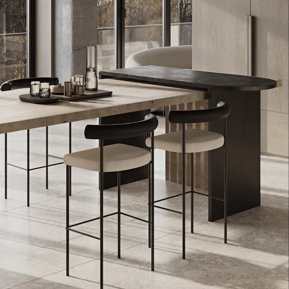 Two tall counter stools with slender matte-black steel frames, beige upholstered seats and curved black faux-leather backrests tucked at a light stone island in a contemporary kitchen setting.