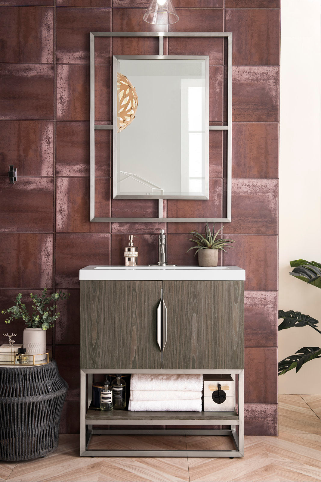 Freestanding modern single-sink bathroom vanity with a white stone top, solid wood stone-gray cabinet, metal base and chrome faucet, framed mirror above, and open lower shelf holding folded towels and bath accessories.