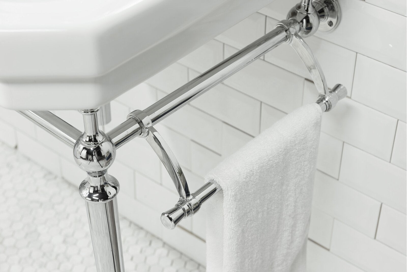 Close-up of a polished chrome metal console sink frame with a white ceramic top and integrated towel bar holding a white towel, set against white subway tile in a modern bathroom vanity.