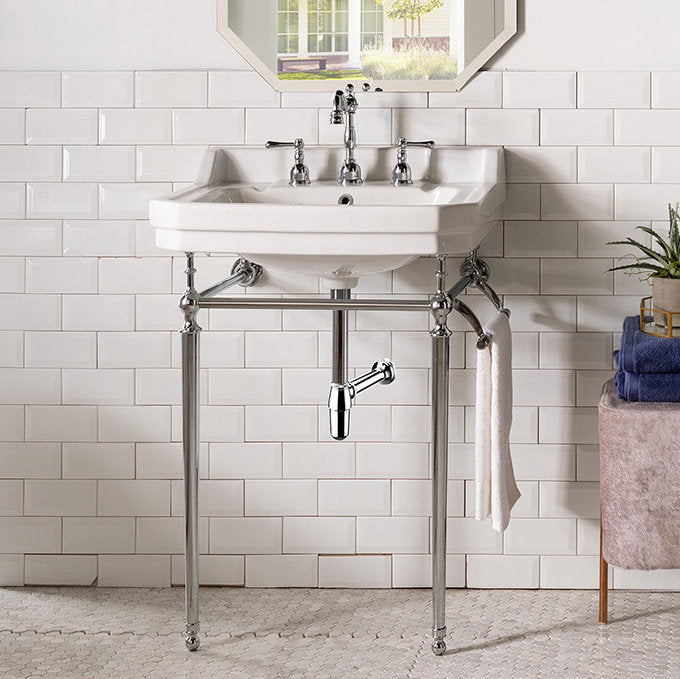 White ceramic single-console bathroom sink with polished chrome metal legs and exposed plumbing, center faucet and towel ring, shown against white subway tile with a small upholstered stool and folded towels to the right.