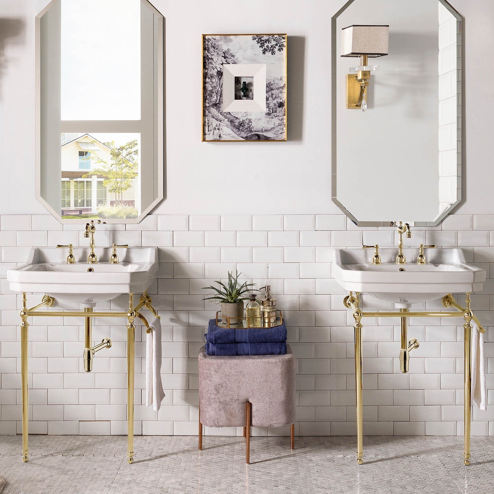 Two white ceramic console sinks with polished brass metal stands and matching brass faucets, mirrored above, flanking a stone-toned upholstered stool stacked with blue towels and bath accessories against white subway tile.