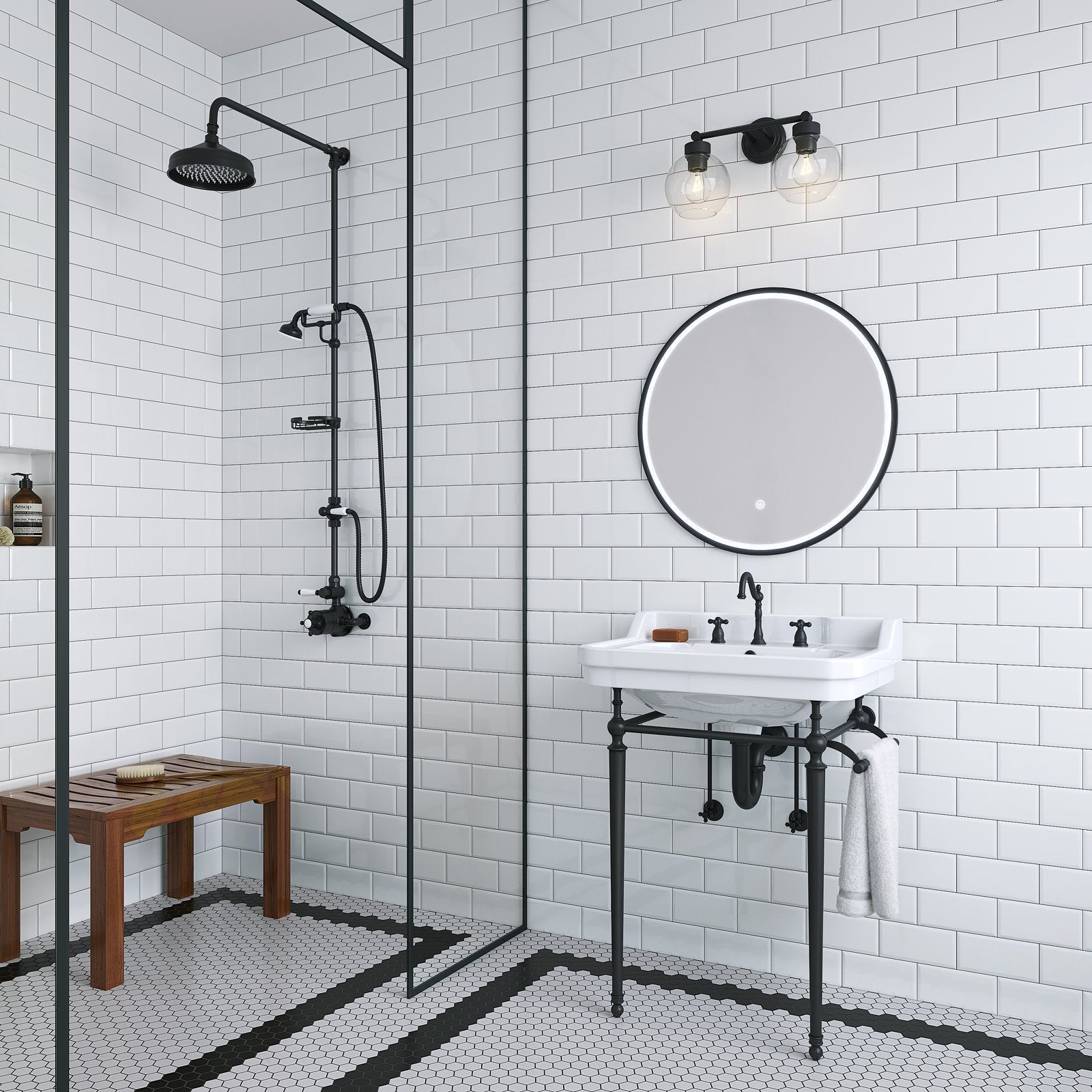 Modern 24-inch white ceramic console sink with stone top and black metal legs, paired with a matte-black faucet and round LED mirror; white subway tile walls, black shower fixtures, hex tile floor and wooden bench.
